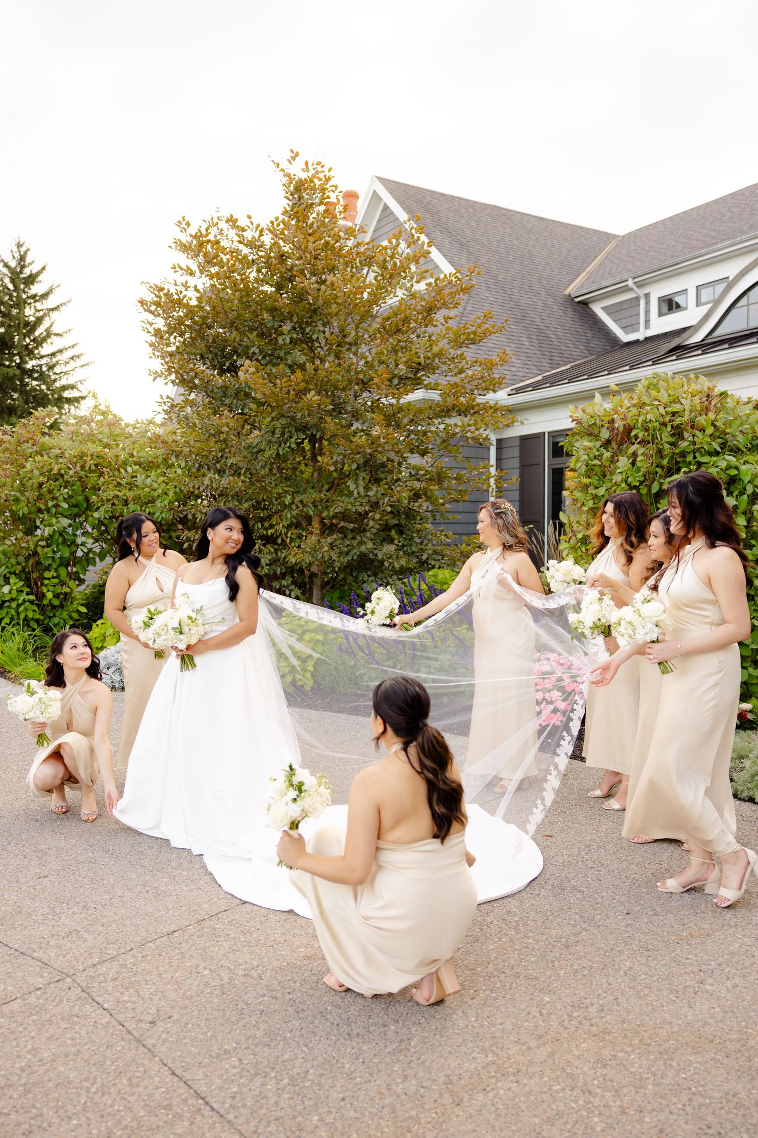 Wedding party in champagne dresses arranged around bride's cathedral veil in outdoor setting.