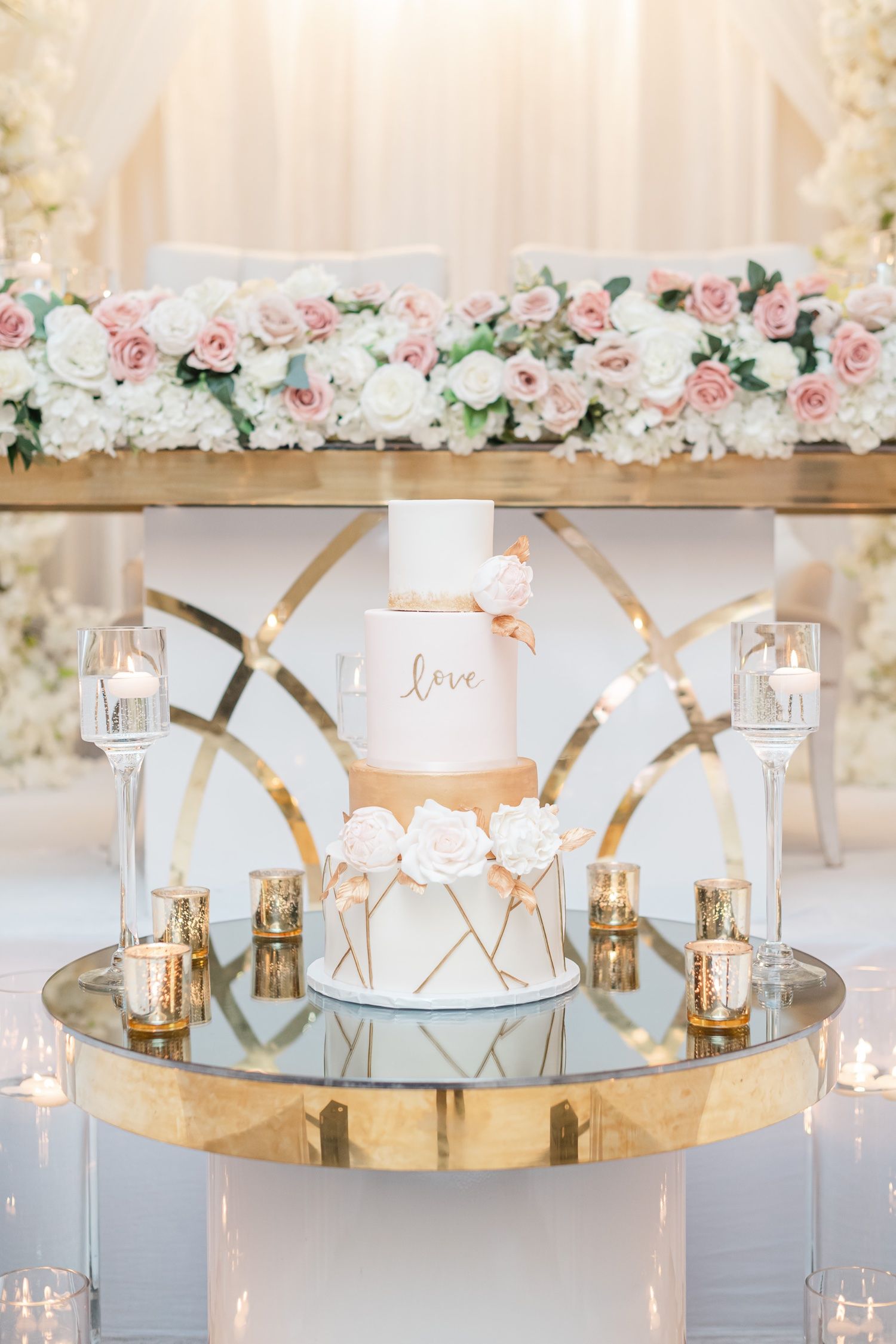 Elegant three-tier white and gold wedding cake decorated with roses on a mirrored gold display stand.