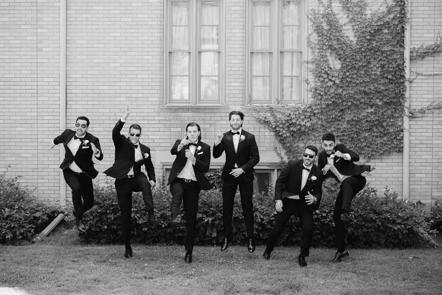 Black and white photo of wedding party jumping in synchronized pose against brick wall.