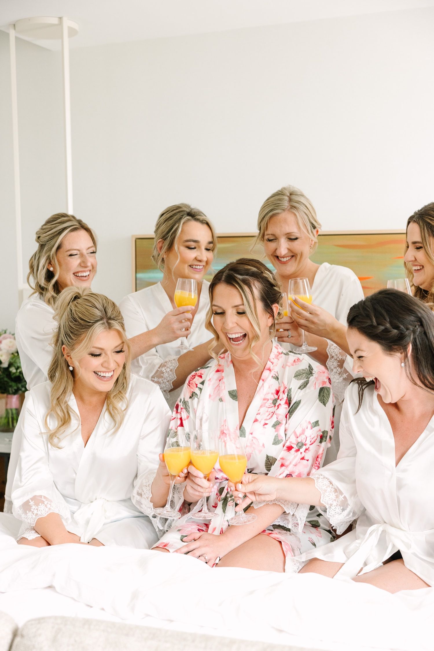 Wedding party in white robes sharing drinks and laughing on bed during preparation.