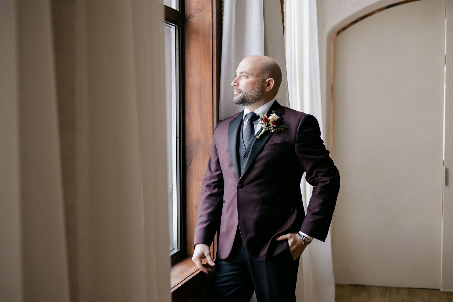 A groom in a burgundy suit jacket gazes thoughtfully out of a window in a classic interior setting.