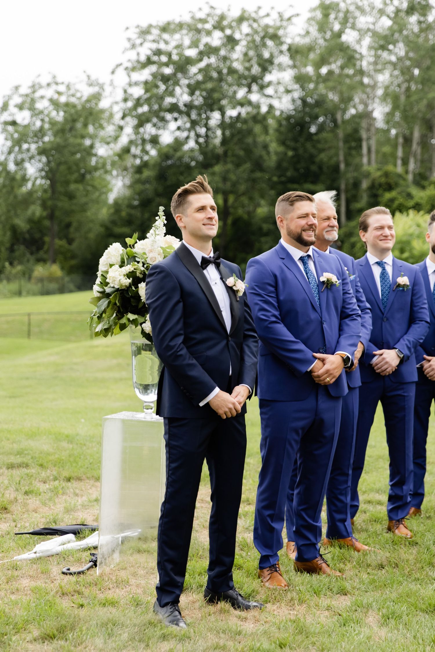 Wedding party in blue and navy suits stands attentively during outdoor ceremony.