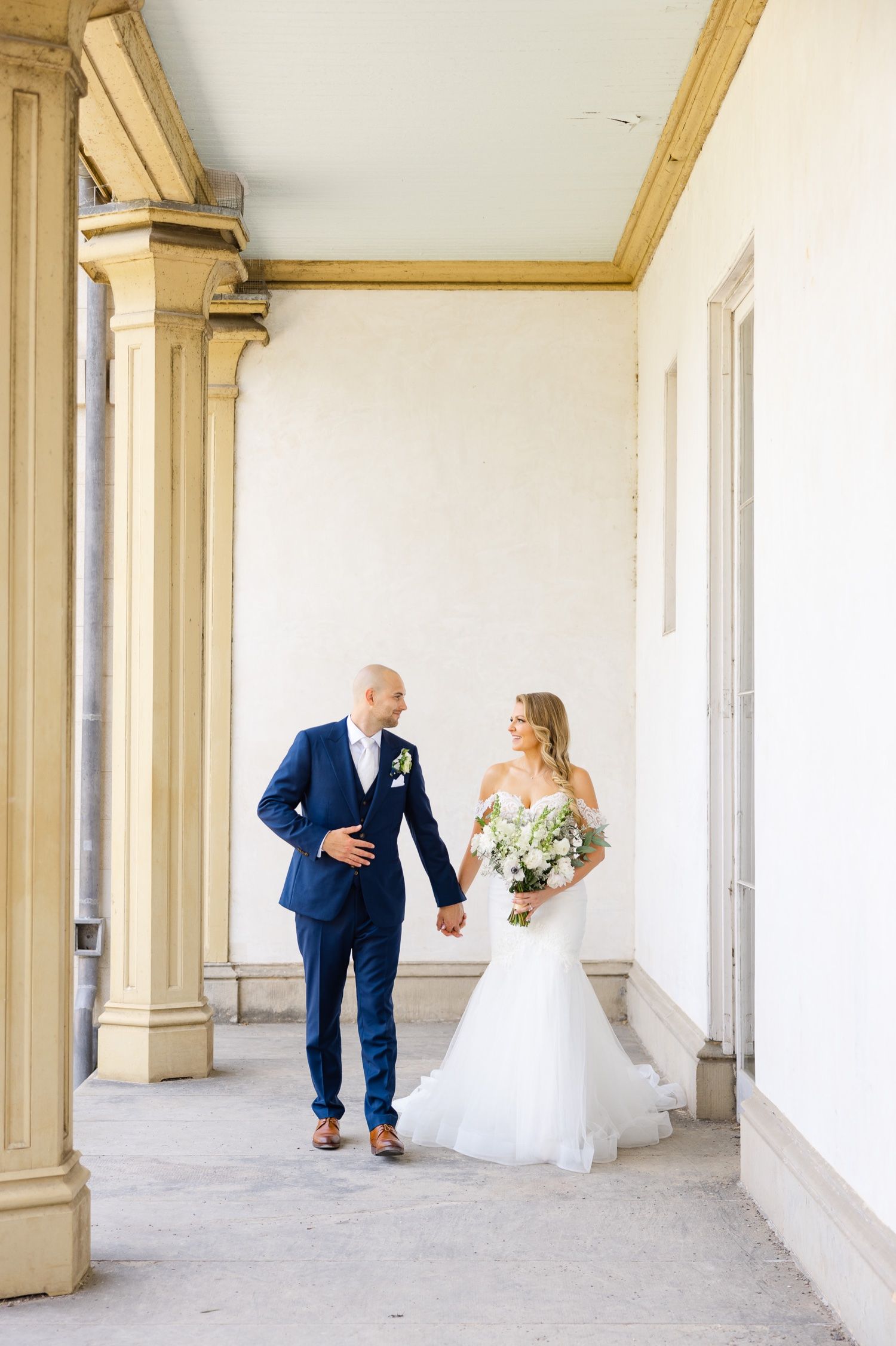 A newlywed couple shares a moment walking along a columned porch with white bouquet.