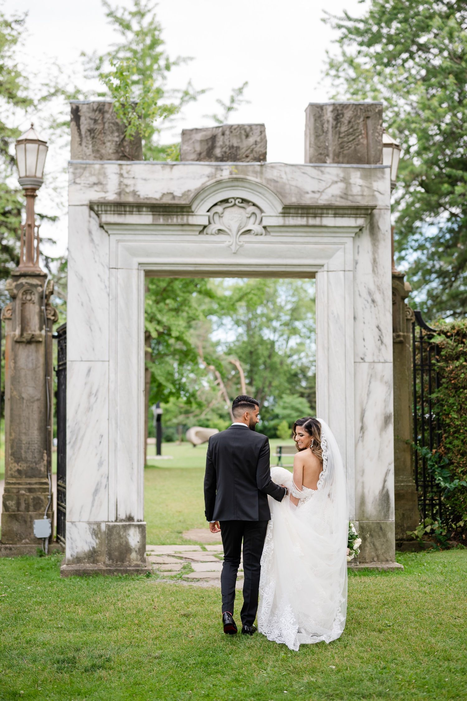 Couple in wedding attire photographed from behind while standing at historic stone archway entrance.