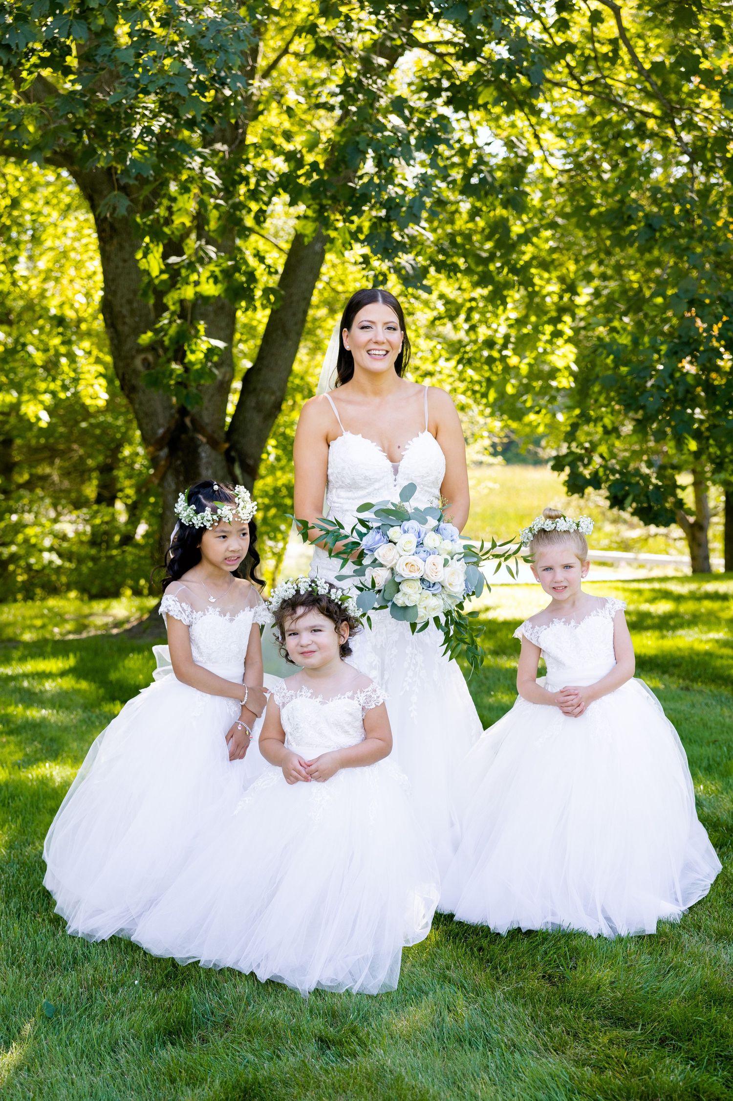 White-gowned figures pose gracefully on lush green grass surrounded by sunlit trees.