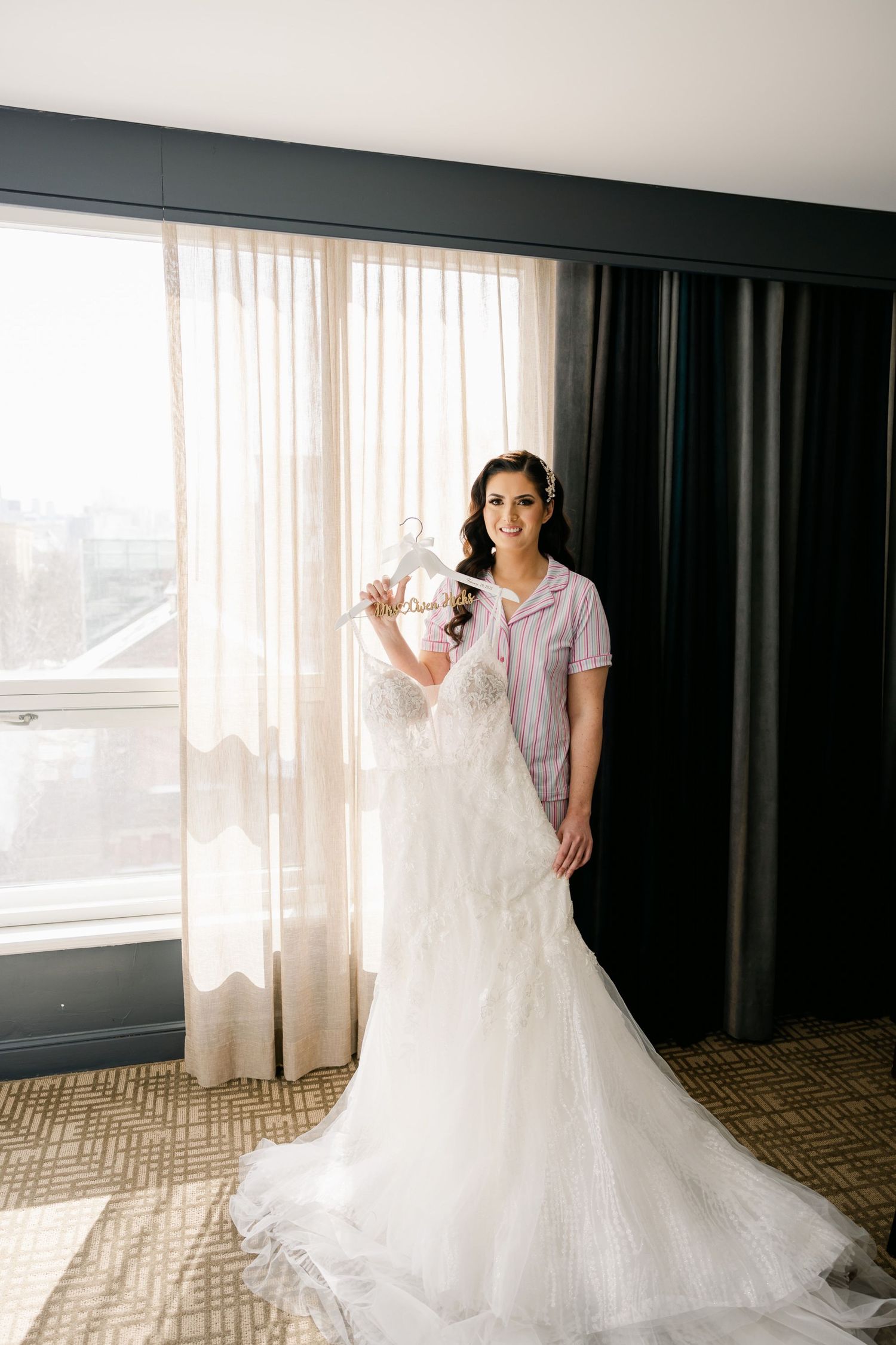 Bride in a pink robe holding her white wedding gown in a sunlit hotel room.