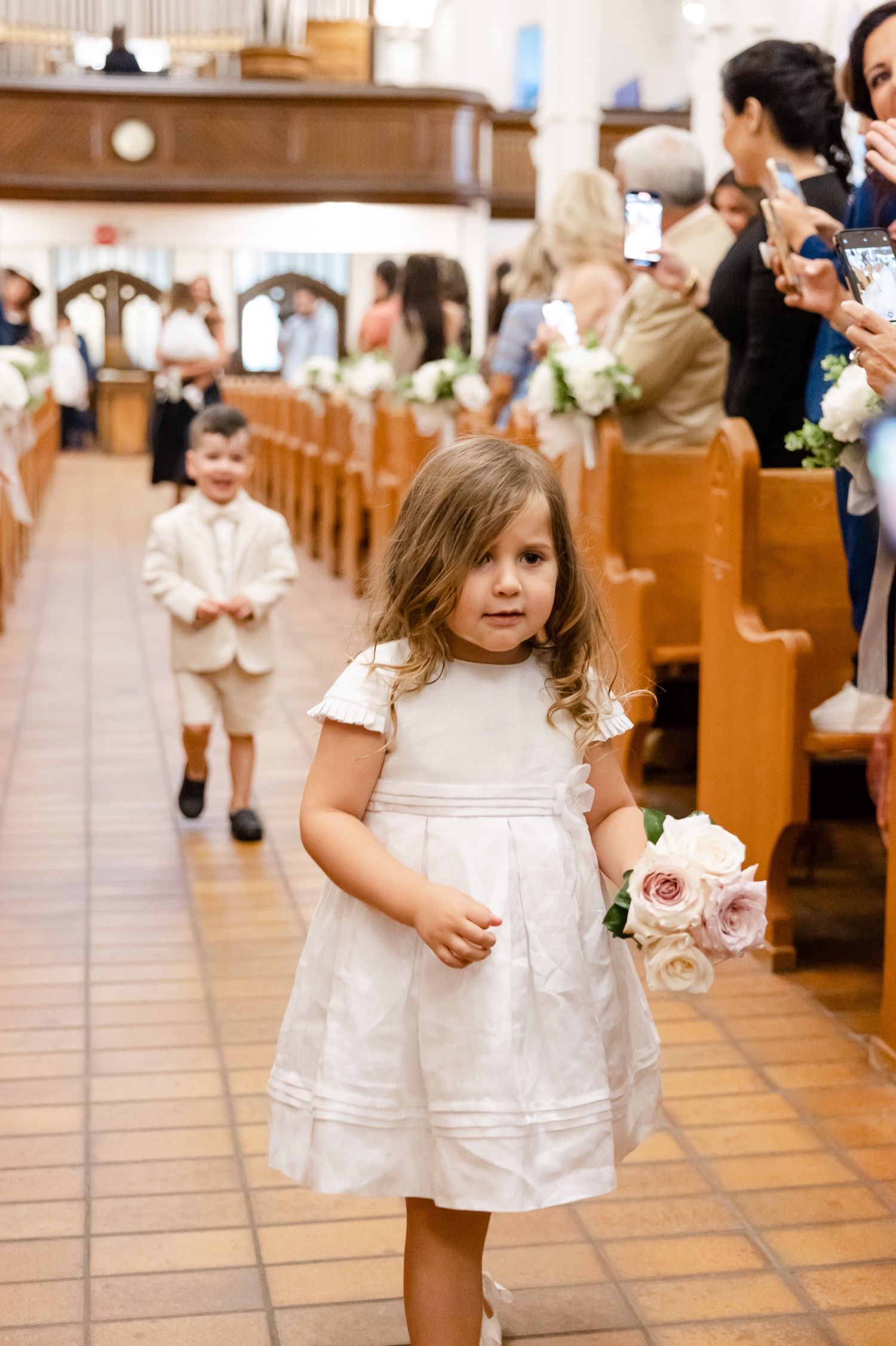 Young flower girl in white dress carrying pink bouquet during church wedding ceremony.