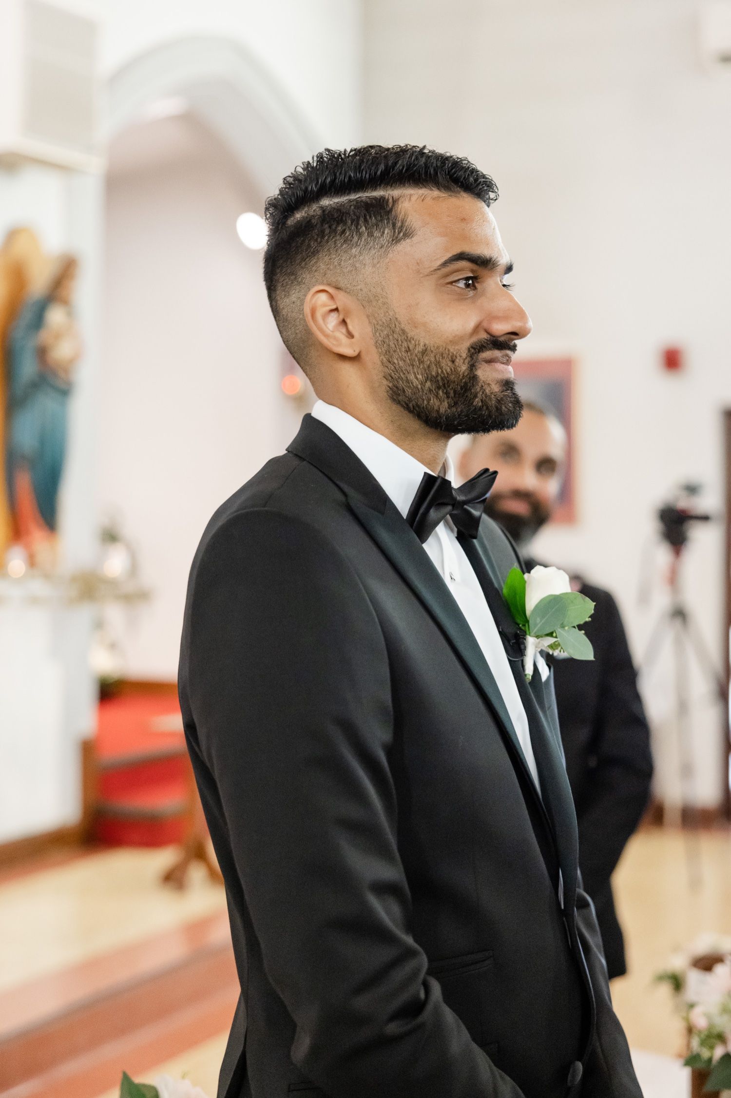 A groom in a classic black tuxedo with a white boutonniere smiles during his wedding ceremony.