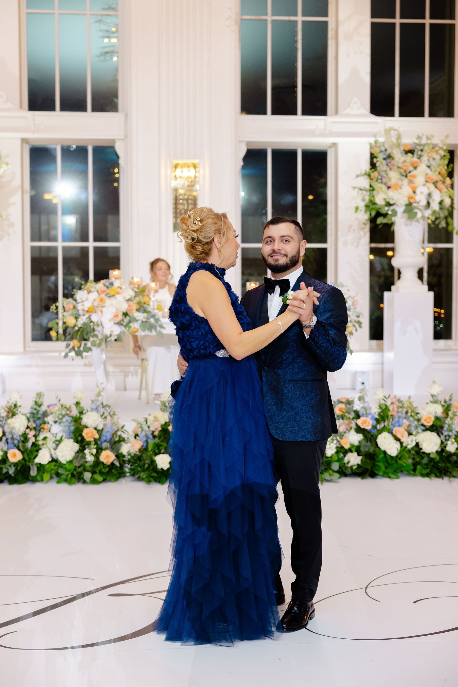 A couple dances together in an elegant white ballroom with blue gown and black tuxedo against floral decorations.