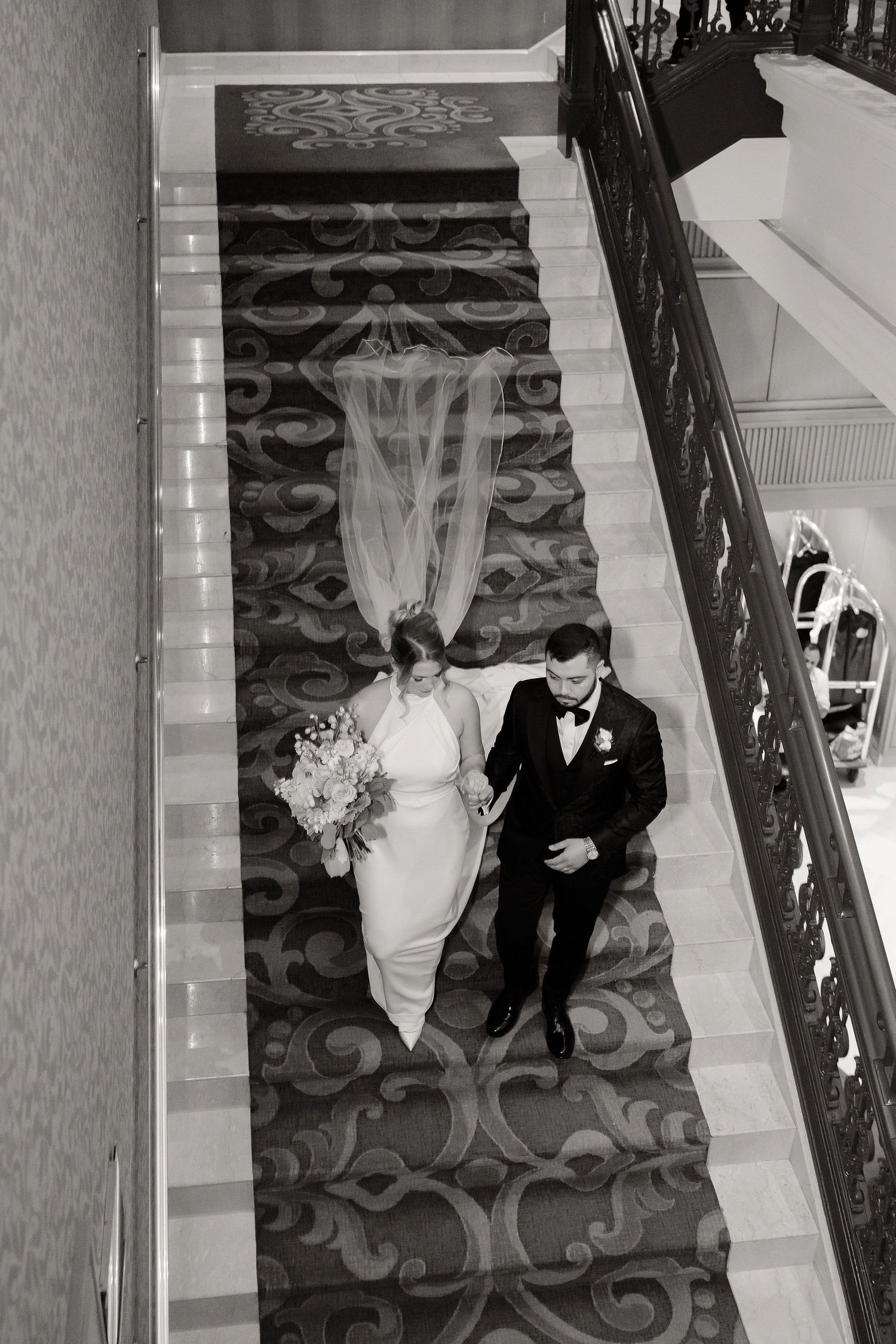 Black and white photo of a couple descending an elegant staircase with patterned carpet.