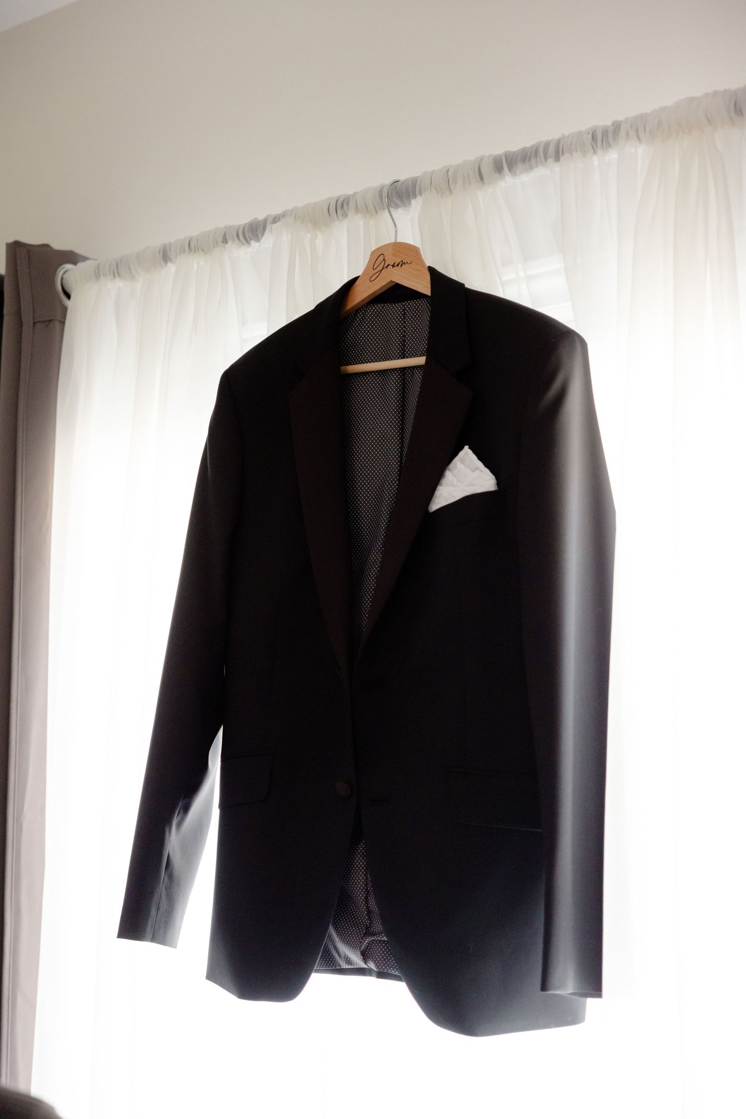 Black formal suit jacket hanging on wooden hanger against white curtain background.