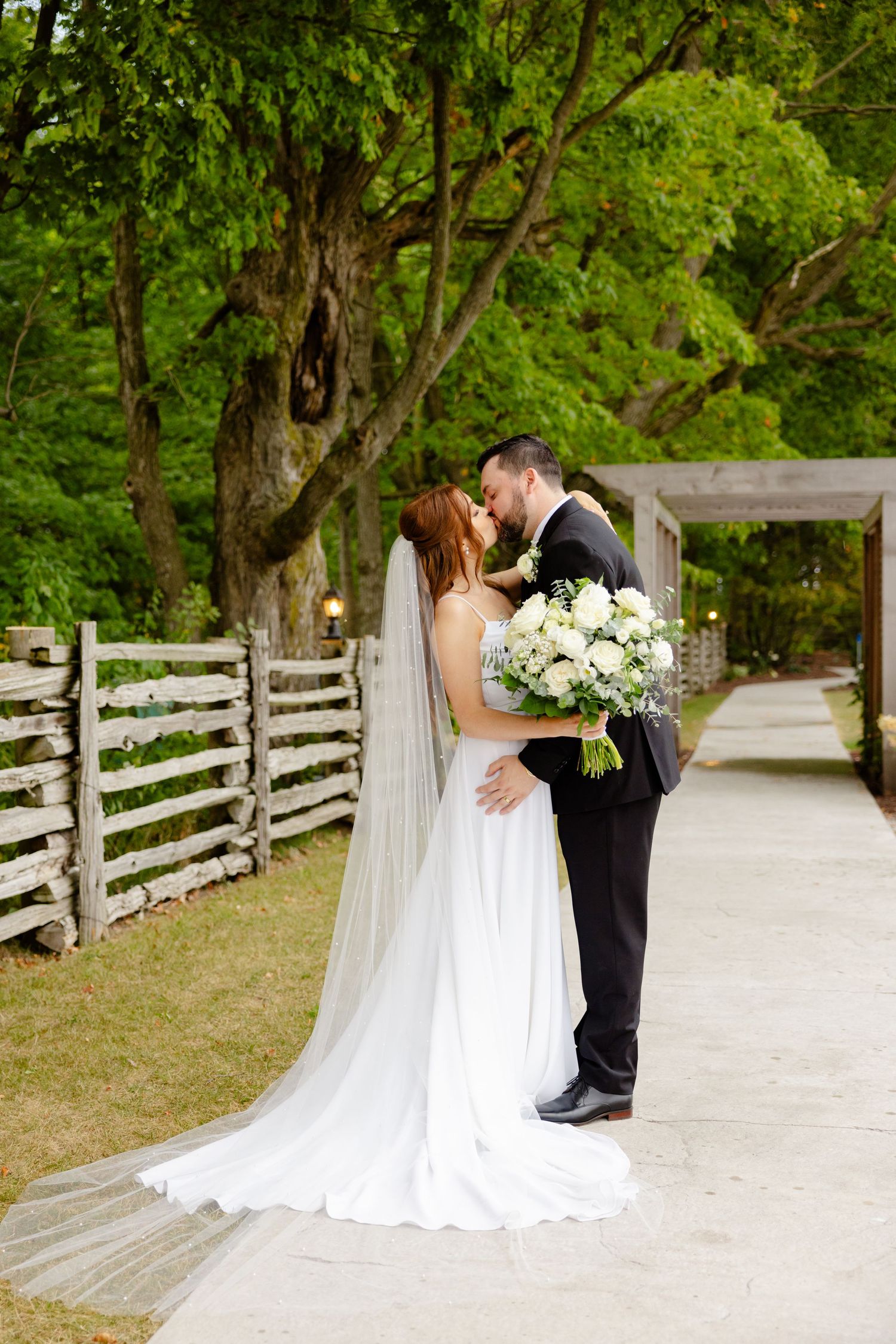 Romantic wedding moment on stone path with rustic fence and green foliage background.