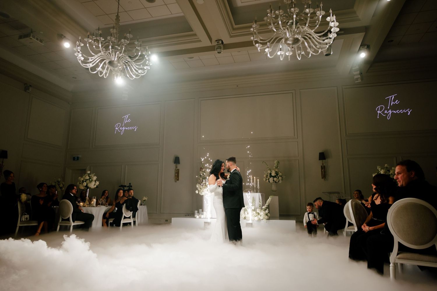 Wedding guests watch a couple's first dance in an elegant ballroom with dramatic lighting.