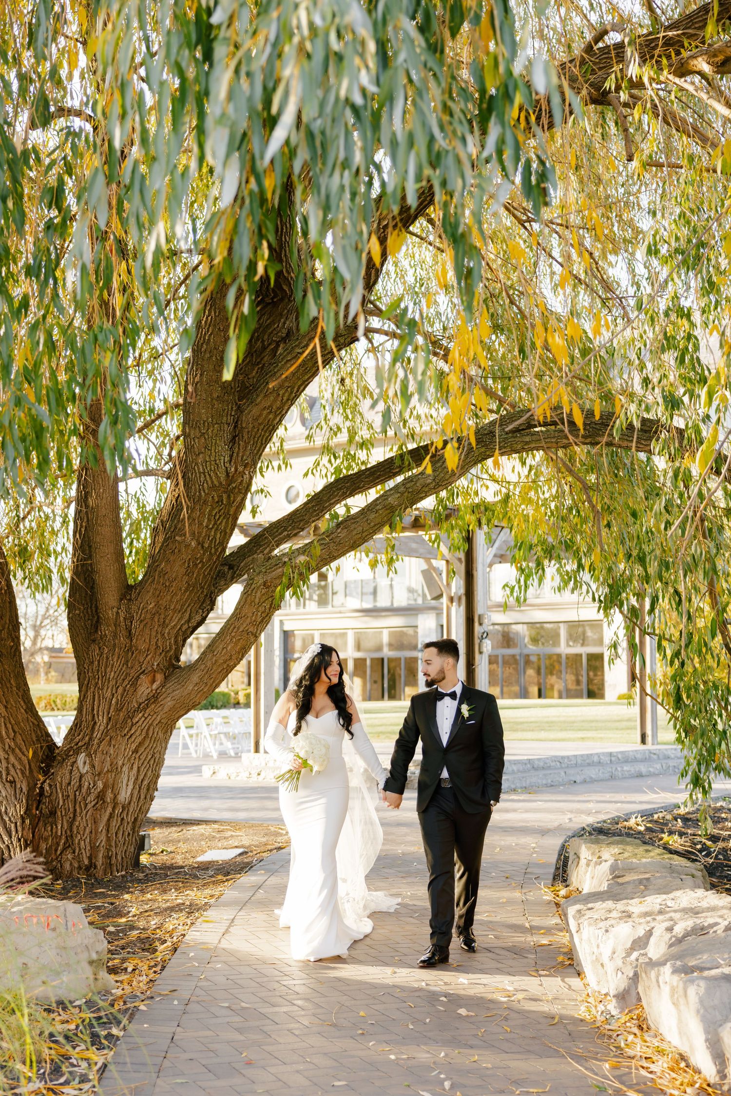 A wedding couple strolling along a stone pathway under autumn willow trees.