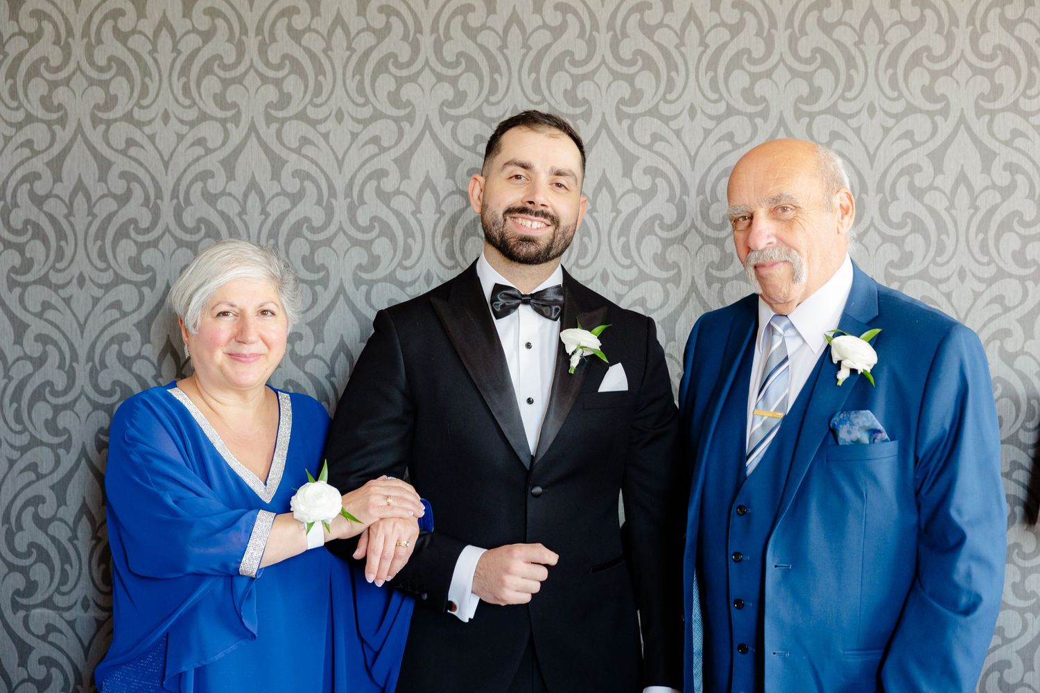 Three people in formal attire smiling together against an elegant grey wallpapered background.
