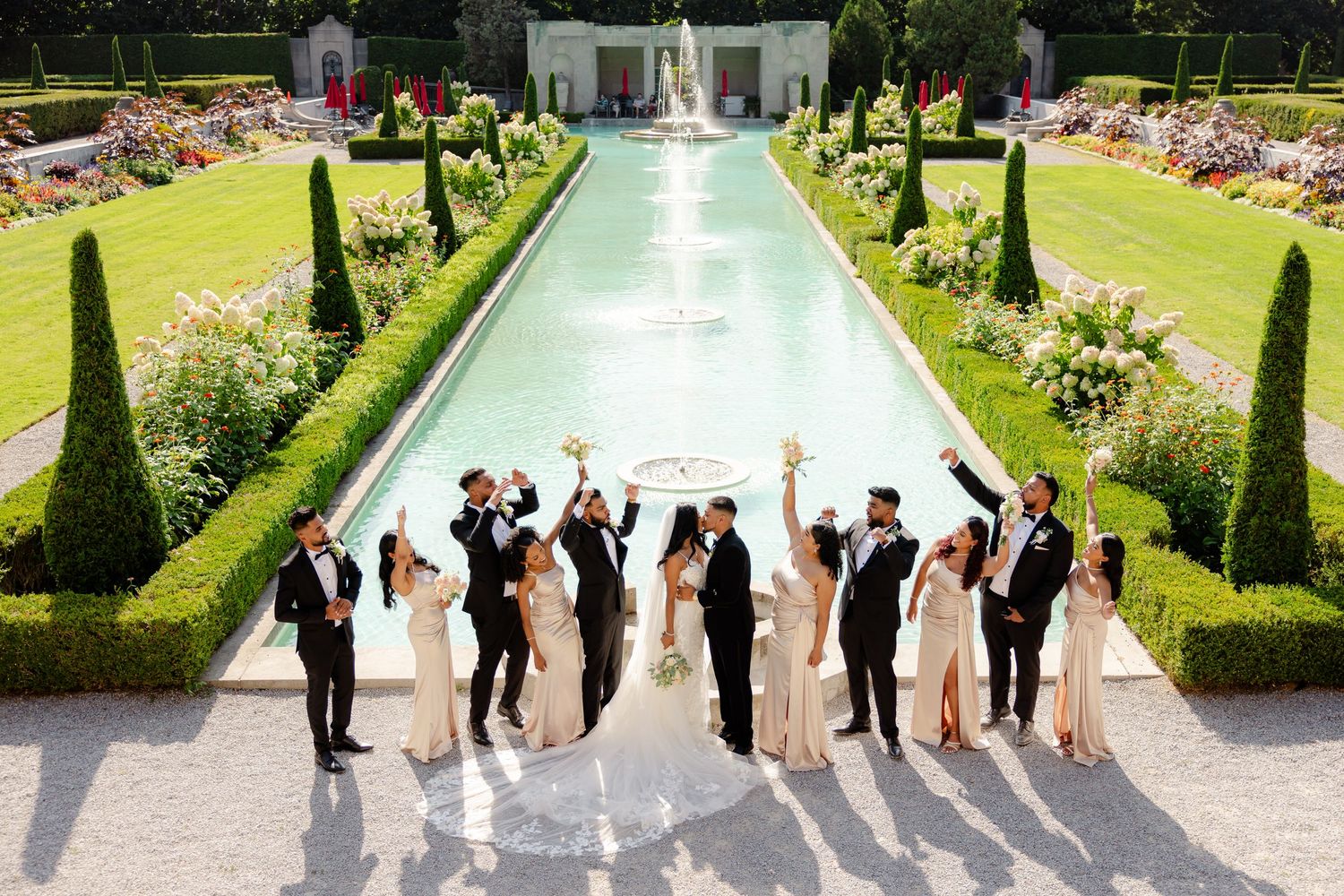 Joyful wedding party celebration pose in front of a grand garden fountain and reflecting pool.
