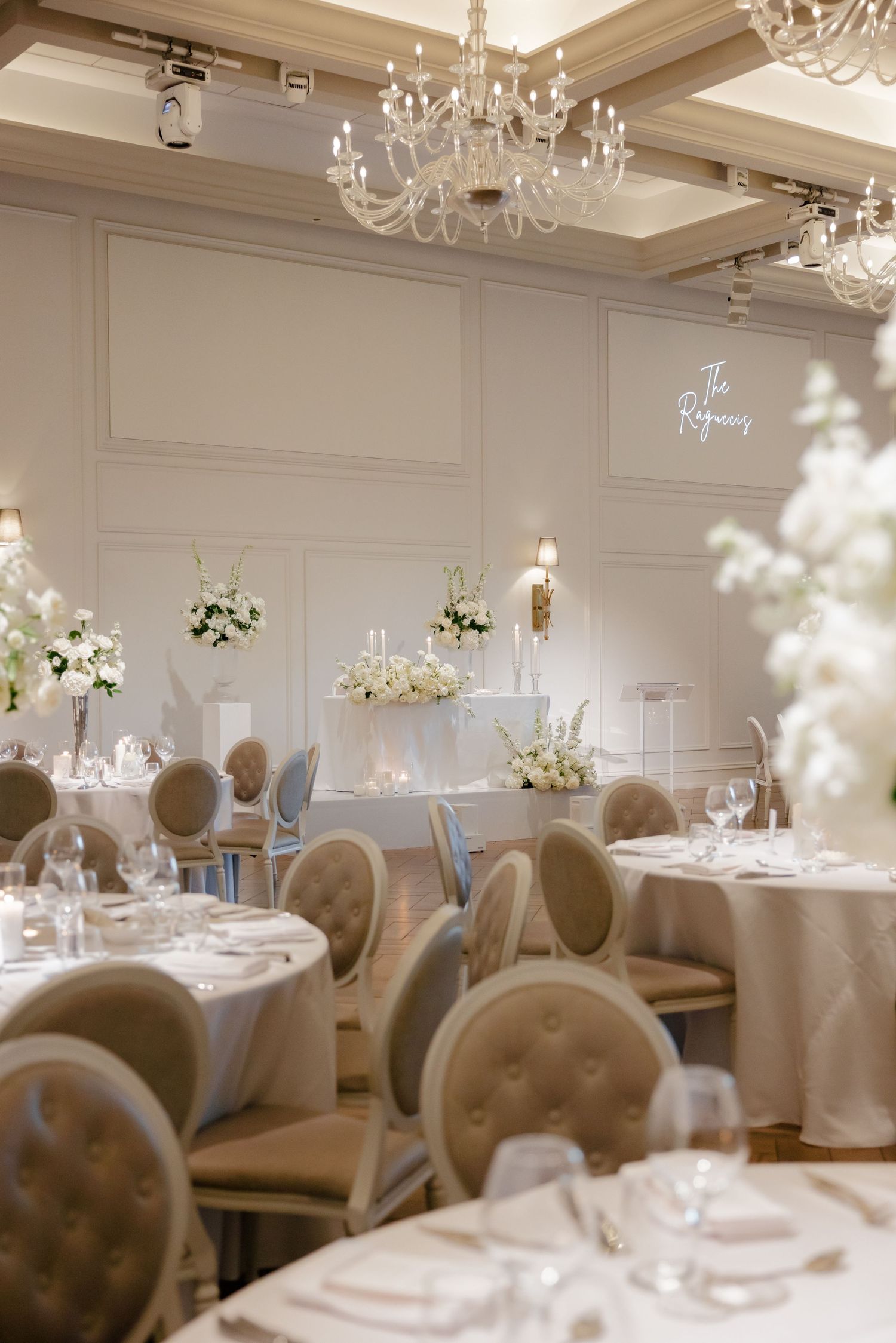 Wedding reception tables with white linens and gray upholstered chairs under ornate crystal chandeliers.