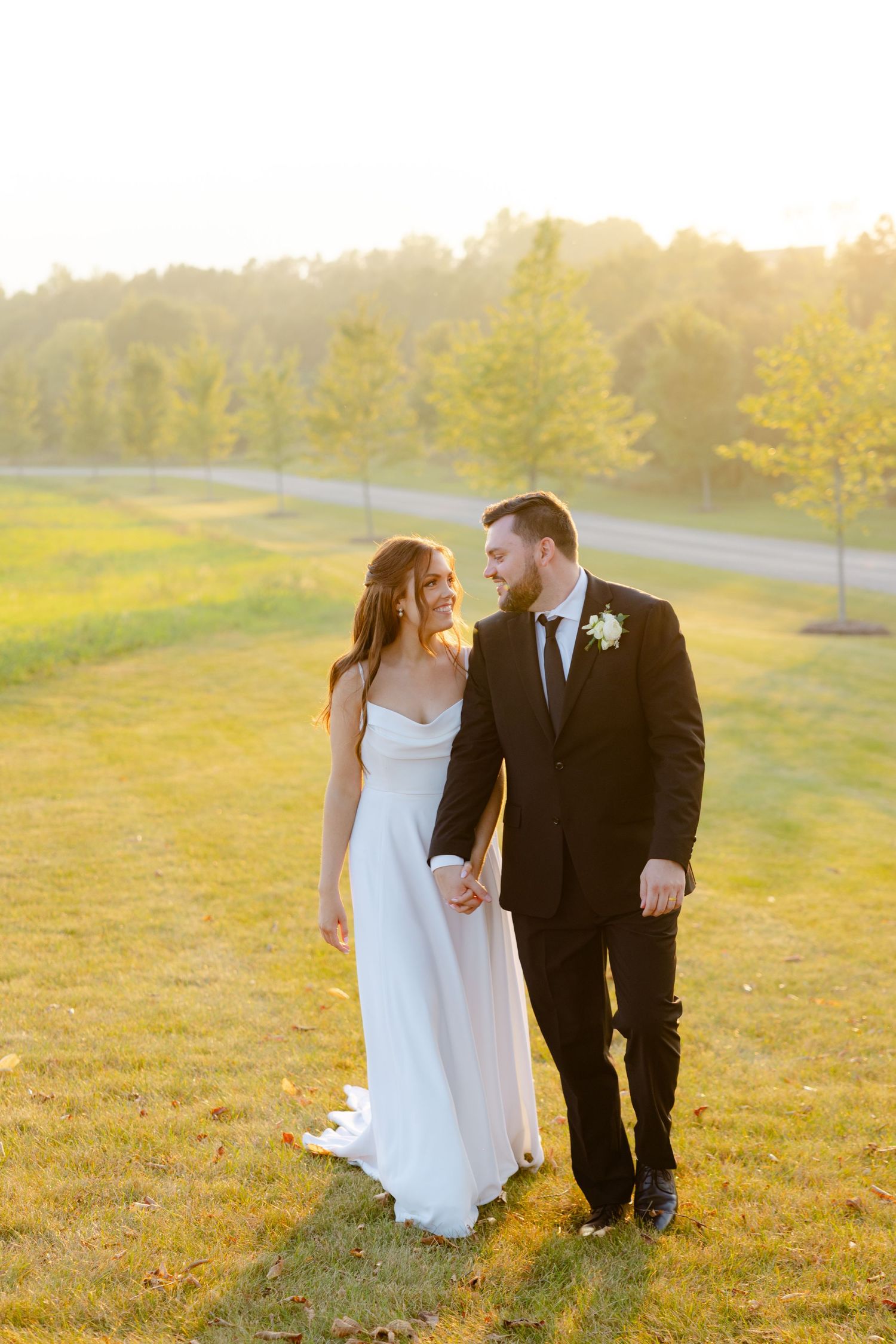 Happy newlyweds smile at each other while standing on a golden-lit hillside during sunset.