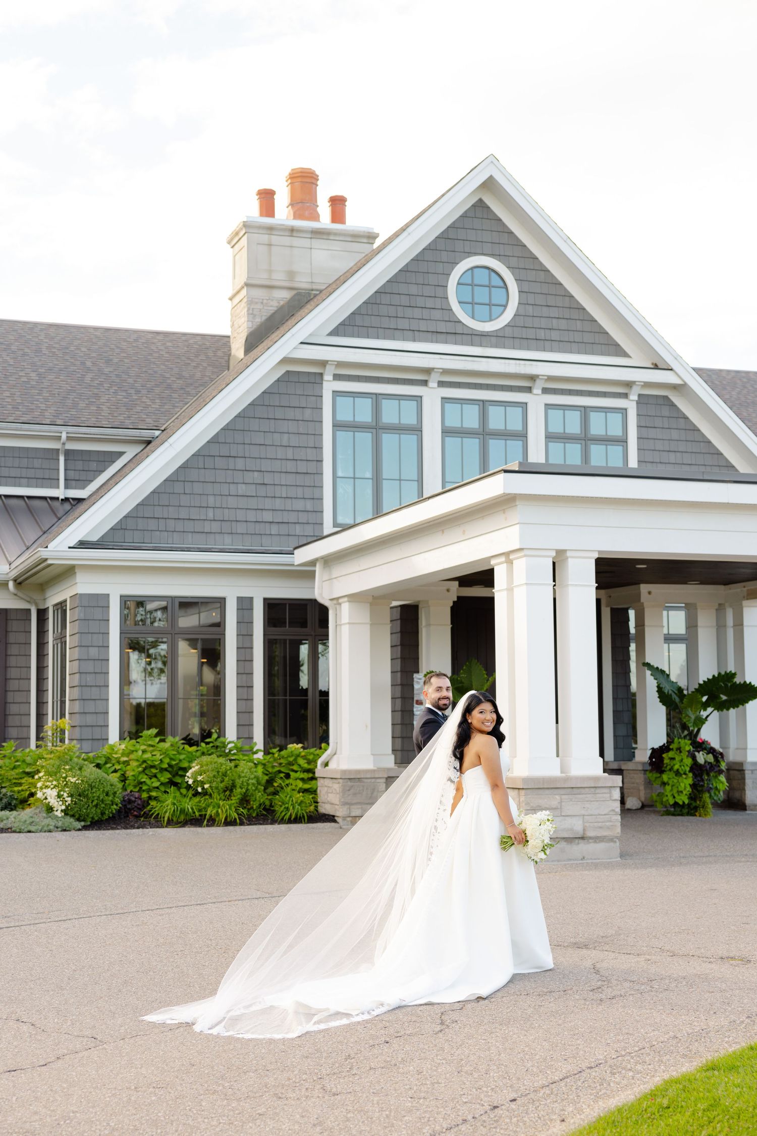 A couple poses in front of a coastal-style home with white trim and gray shingles.