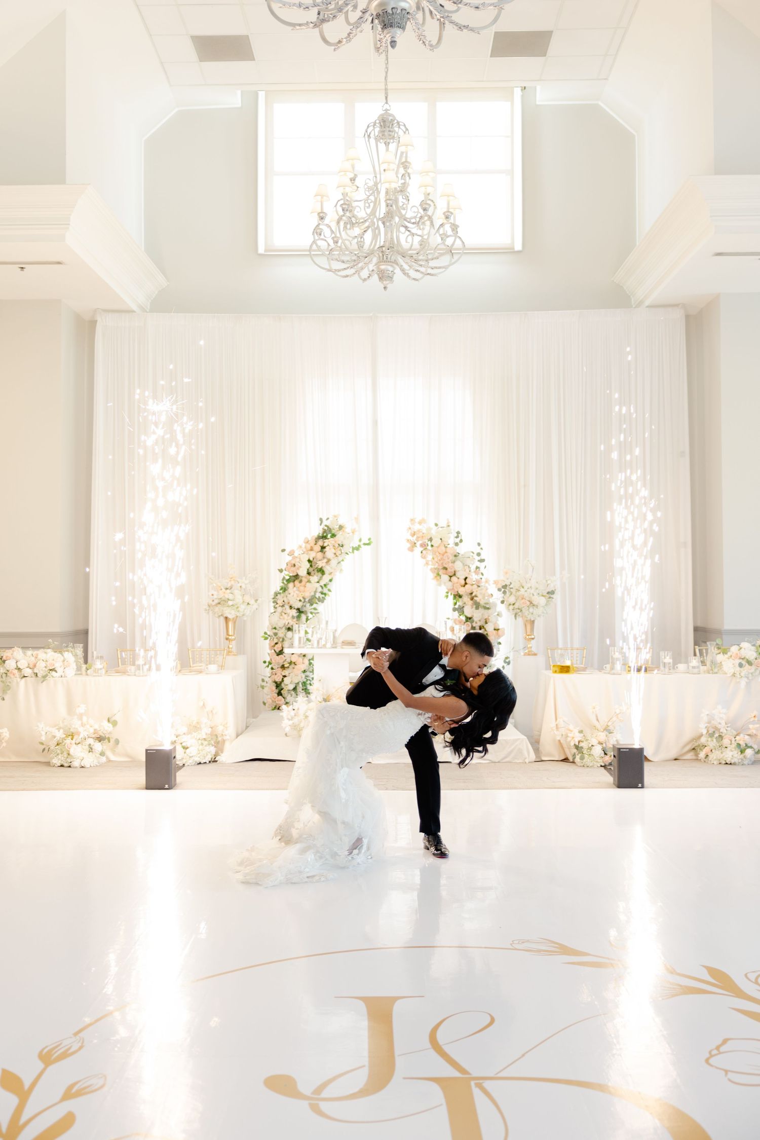 Romantic dance dip pose captured in bright white wedding ballroom with floral arch backdrop.