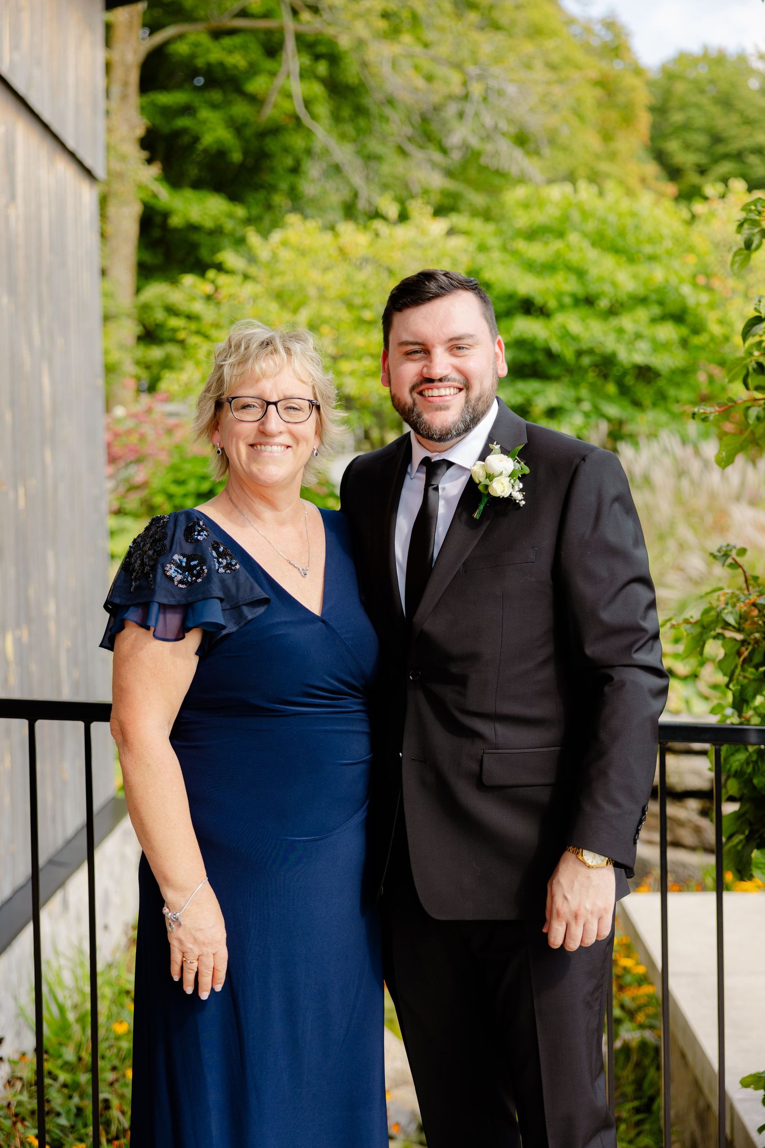 Wedding photo of pair in formal attire standing on wooden deck with greenery backdrop.