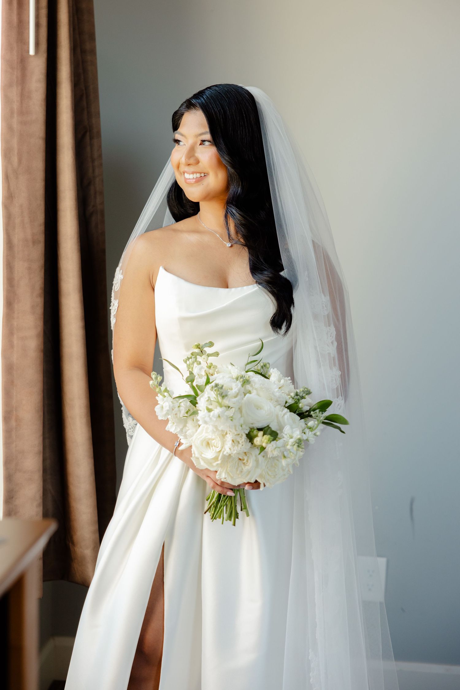 Radiant bride in strapless wedding gown poses with white floral bouquet near window.