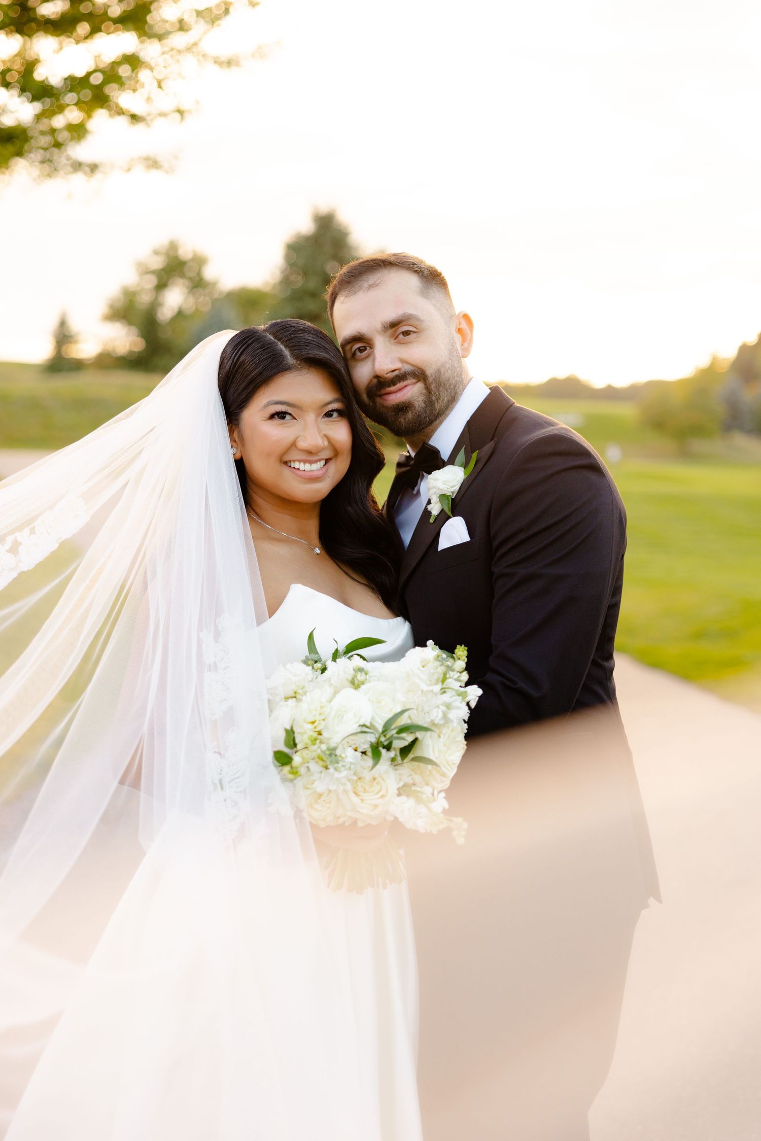 A sun-drenched wedding portrait of a happy couple with flowing veil and white bouquet on a golf course path.