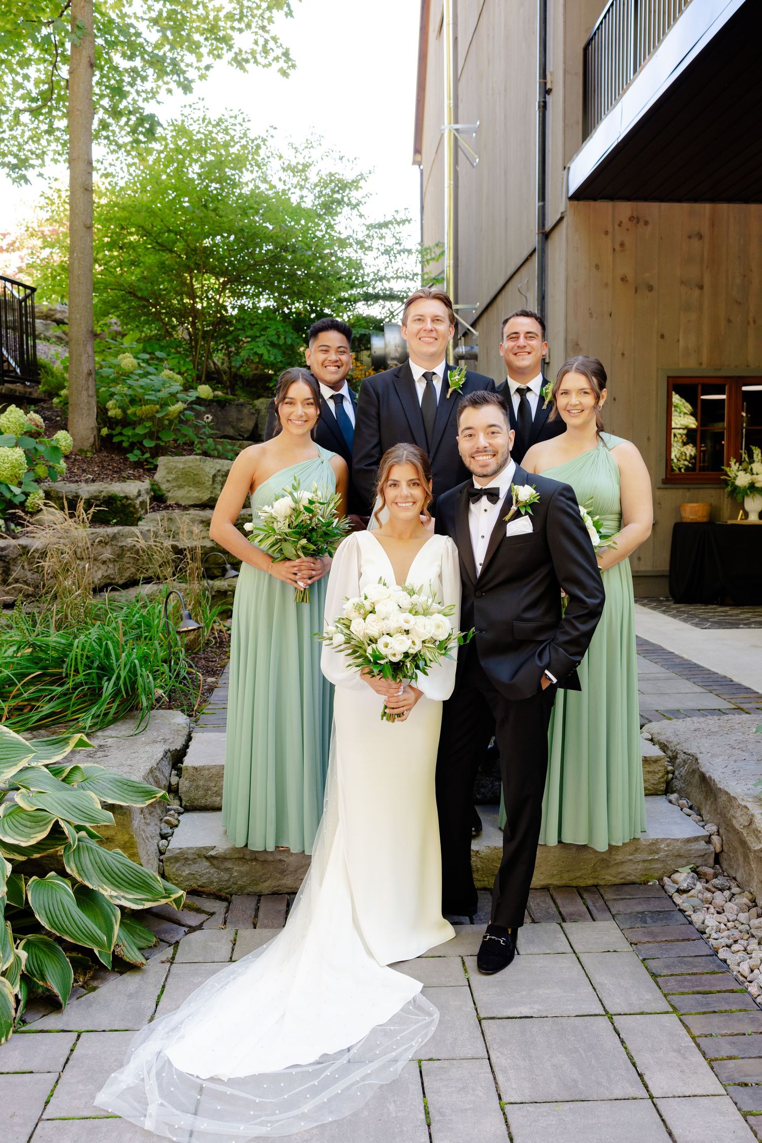 Wedding party in sage green dresses and dark suits pose together on stone patio with landscaping.