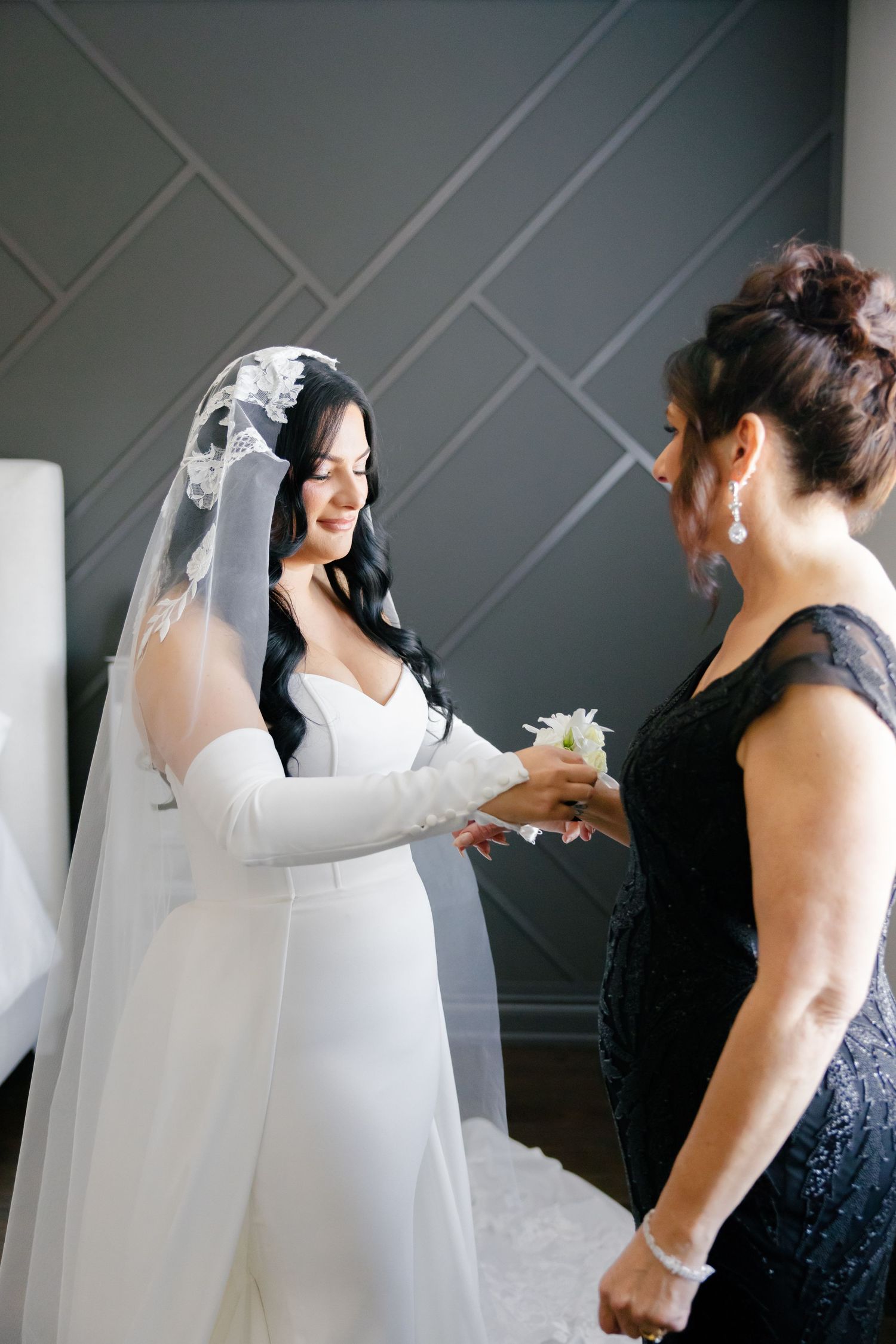A bride in a white dress and veil shares a touching moment with a woman in a navy gown against a geometric wall.