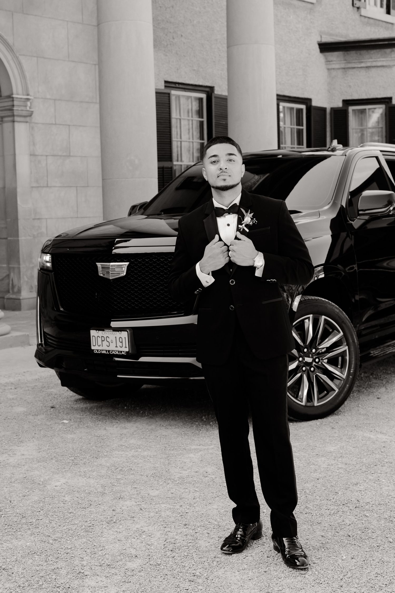 A groom in a black tuxedo adjusts his bowtie while standing in front of a luxury Cadillac SUV and white pillars.