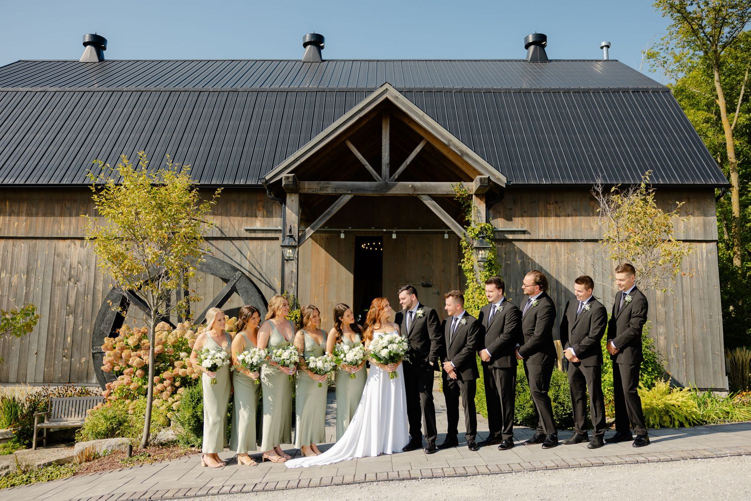 Formal wedding party portrait in front of wooden barn featuring sage green dresses and dark suits.