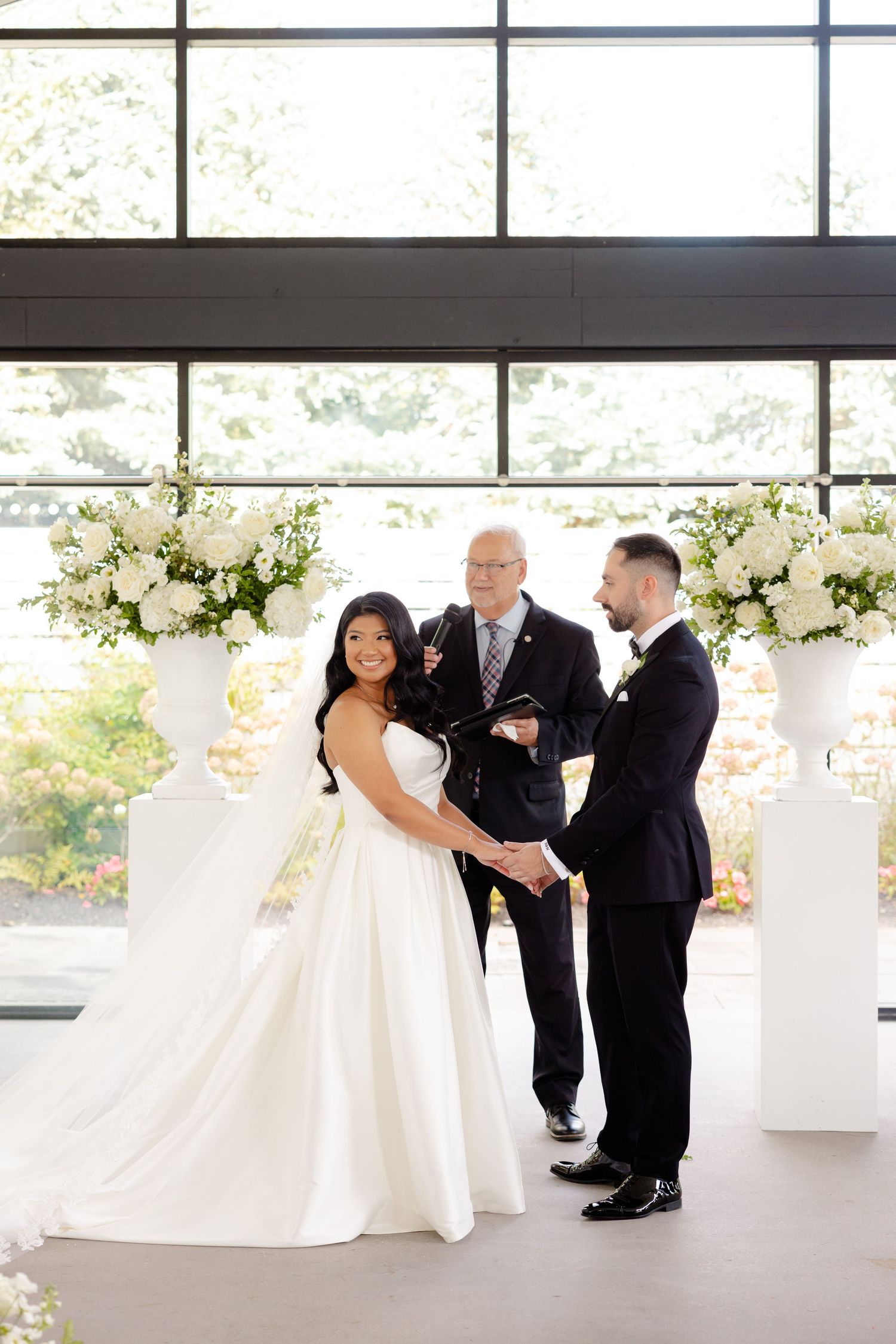 Wedding ceremony in bright indoor venue with white floral arrangements and large windows.