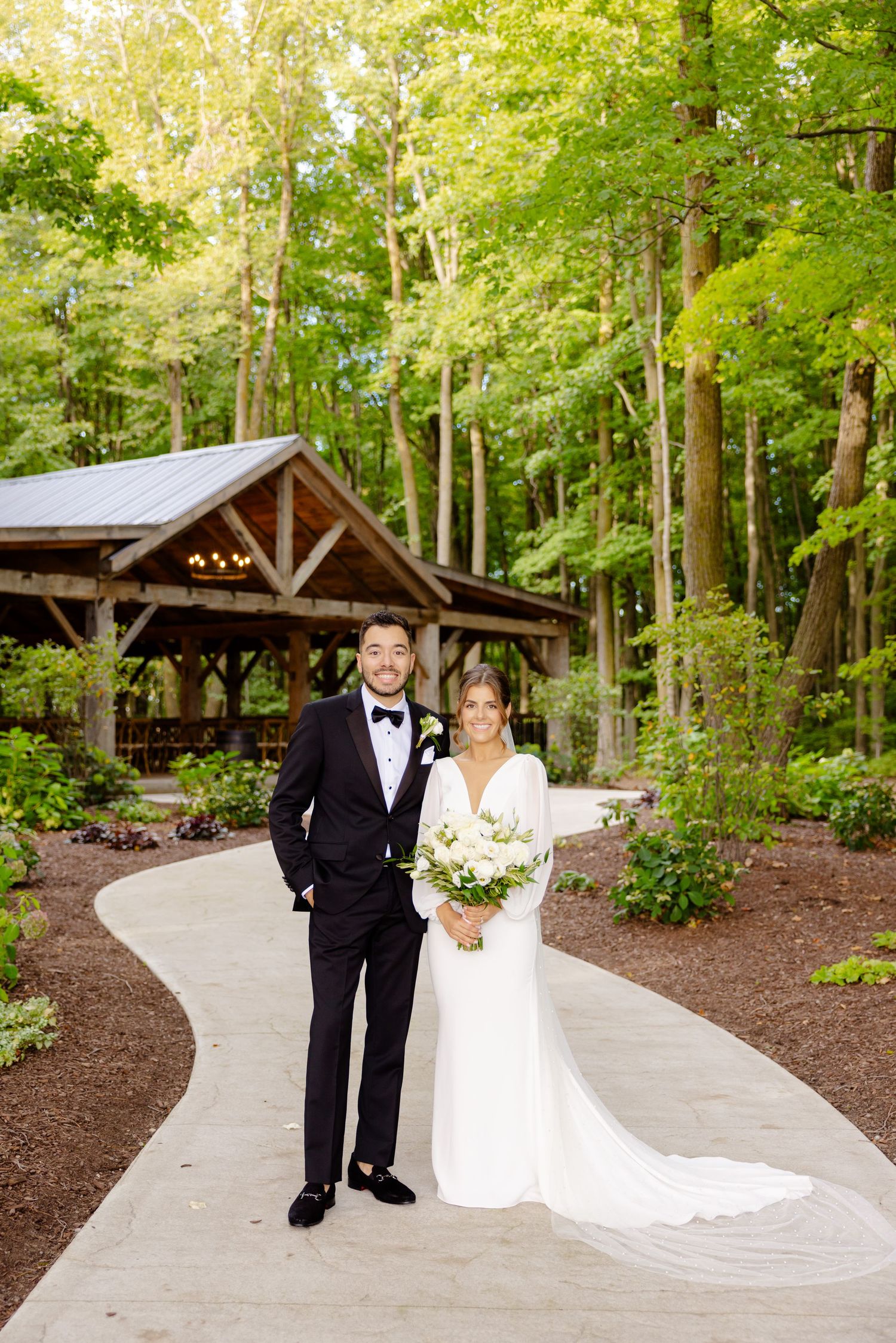 Newlyweds pose on winding concrete path with rustic wooden pavilion and green forest.