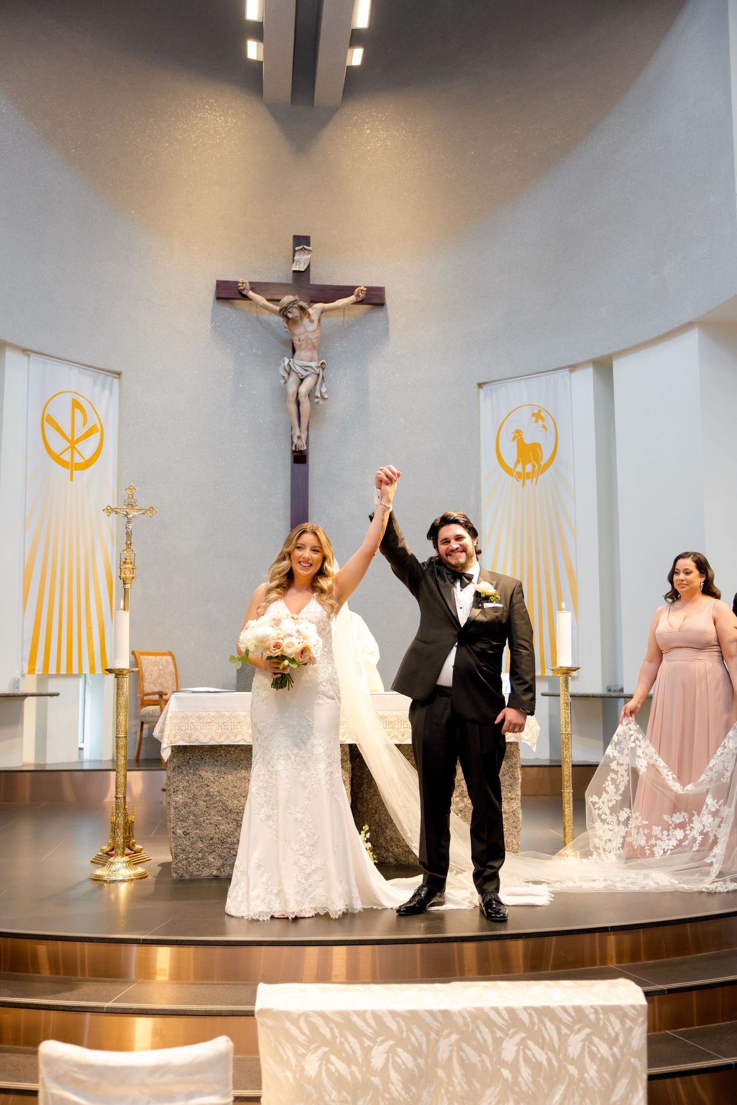 Newlyweds celebrating their marriage with a triumphant pose at the altar of a Catholic church with decorative banners.