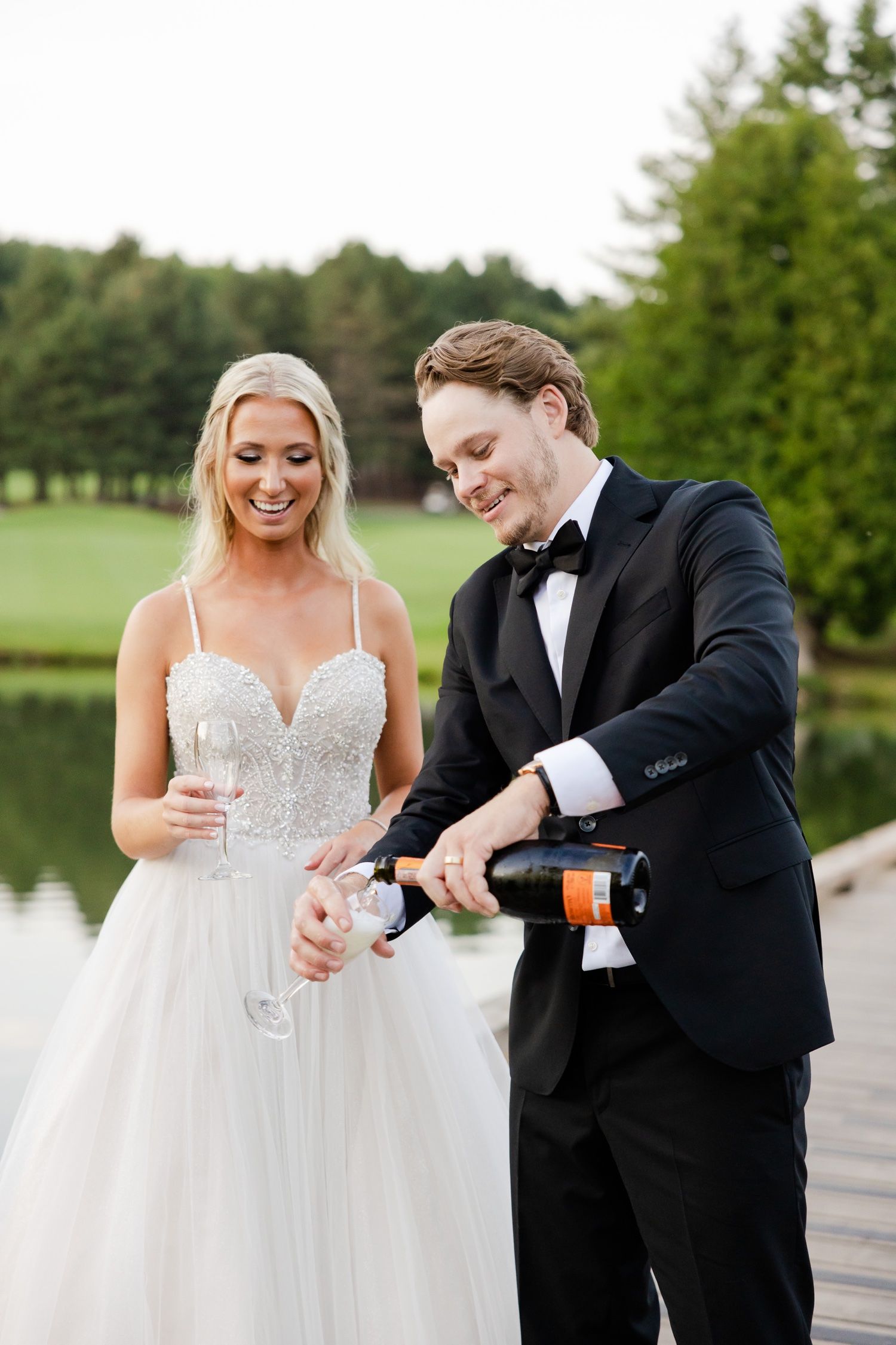 A couple opens champagne during their wedding celebration on a wooden dock with a lake view.