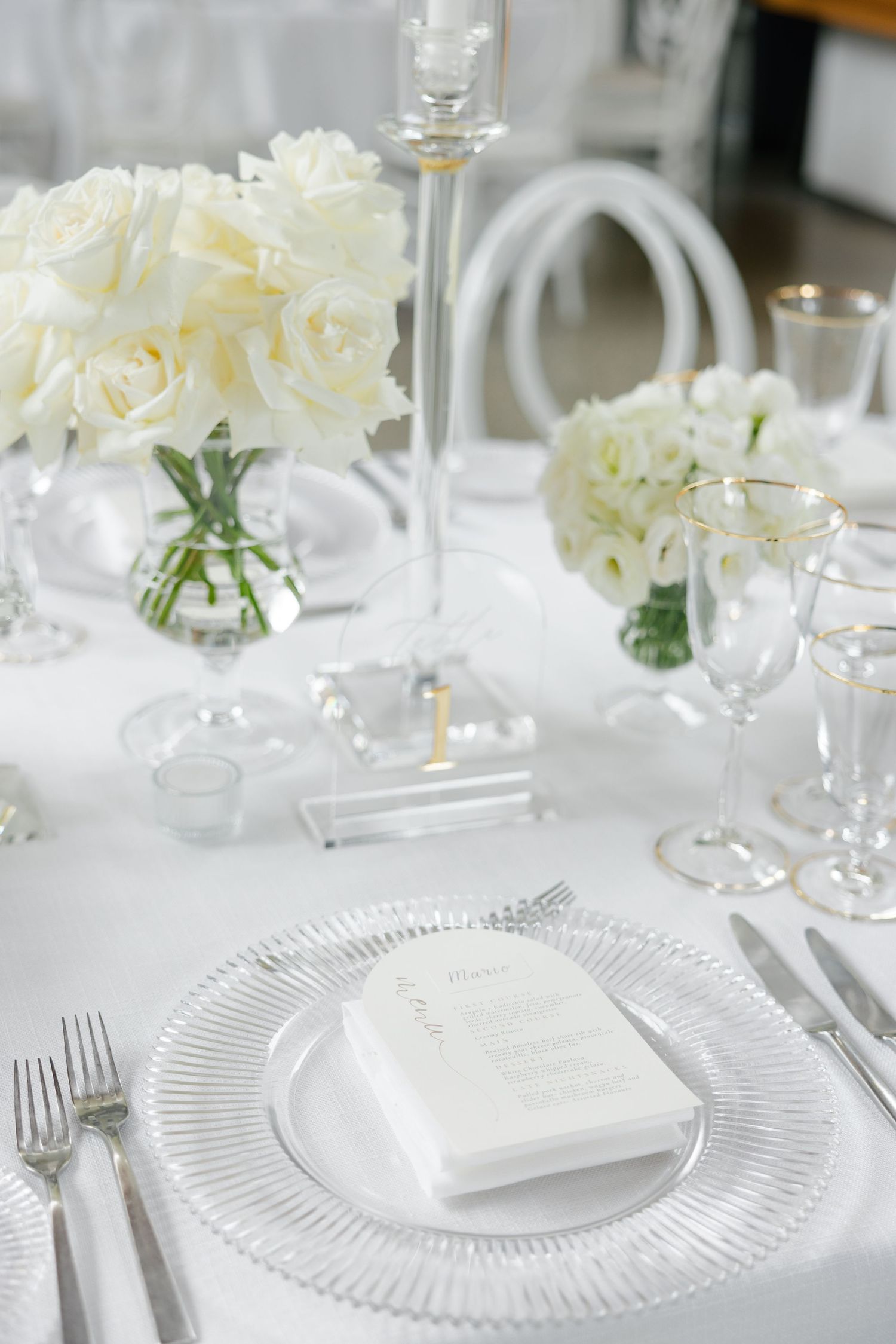 Elegant white place setting with crystal glassware and white roses in a clear vase on a pristine tablecloth.