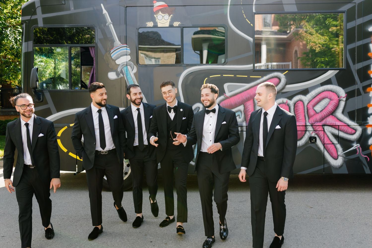 Wedding party in black suits walking together in front of graffiti-decorated party bus.