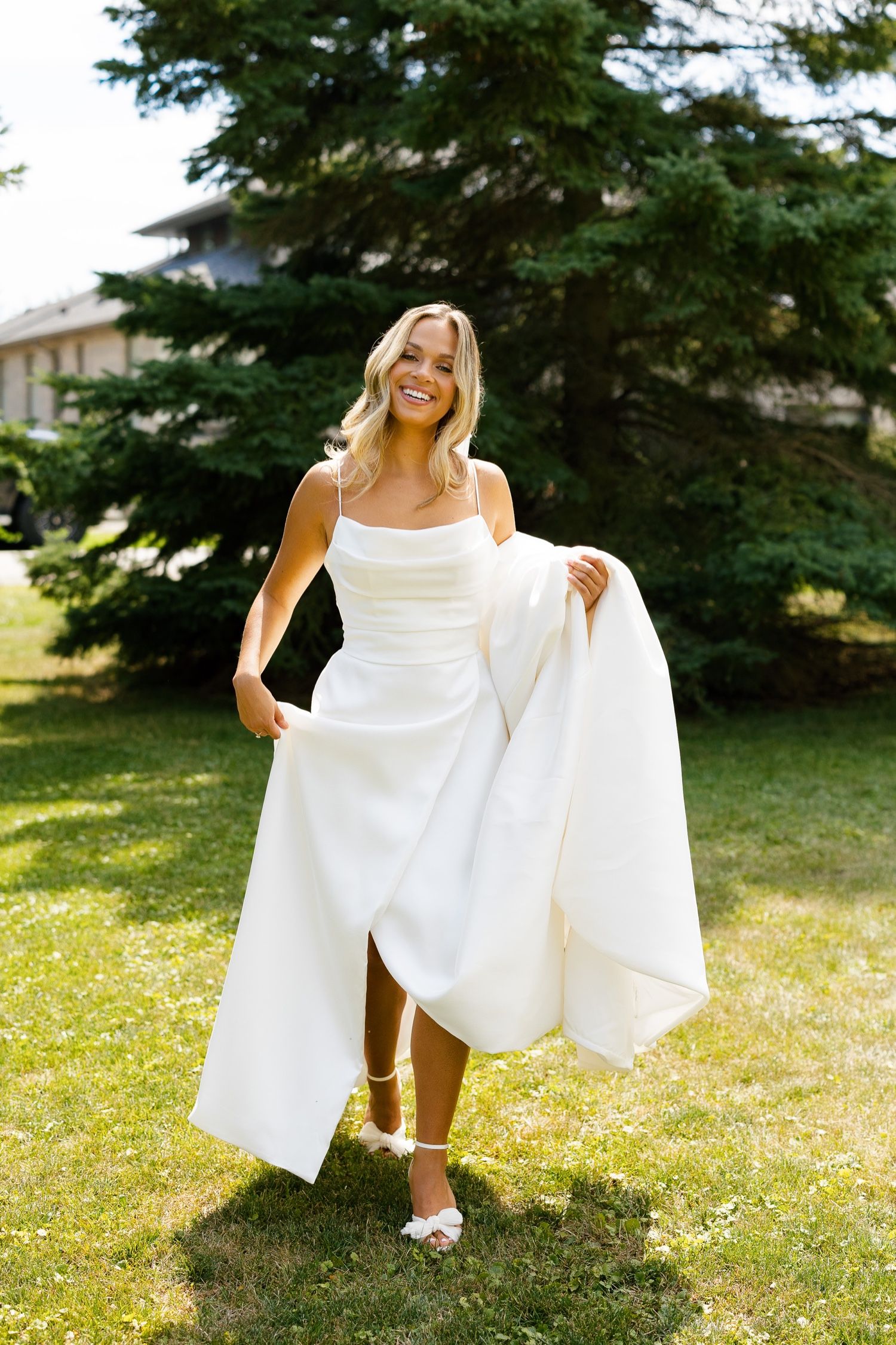 Bride in white dress playfully lifts her gown's skirt while standing on grass.