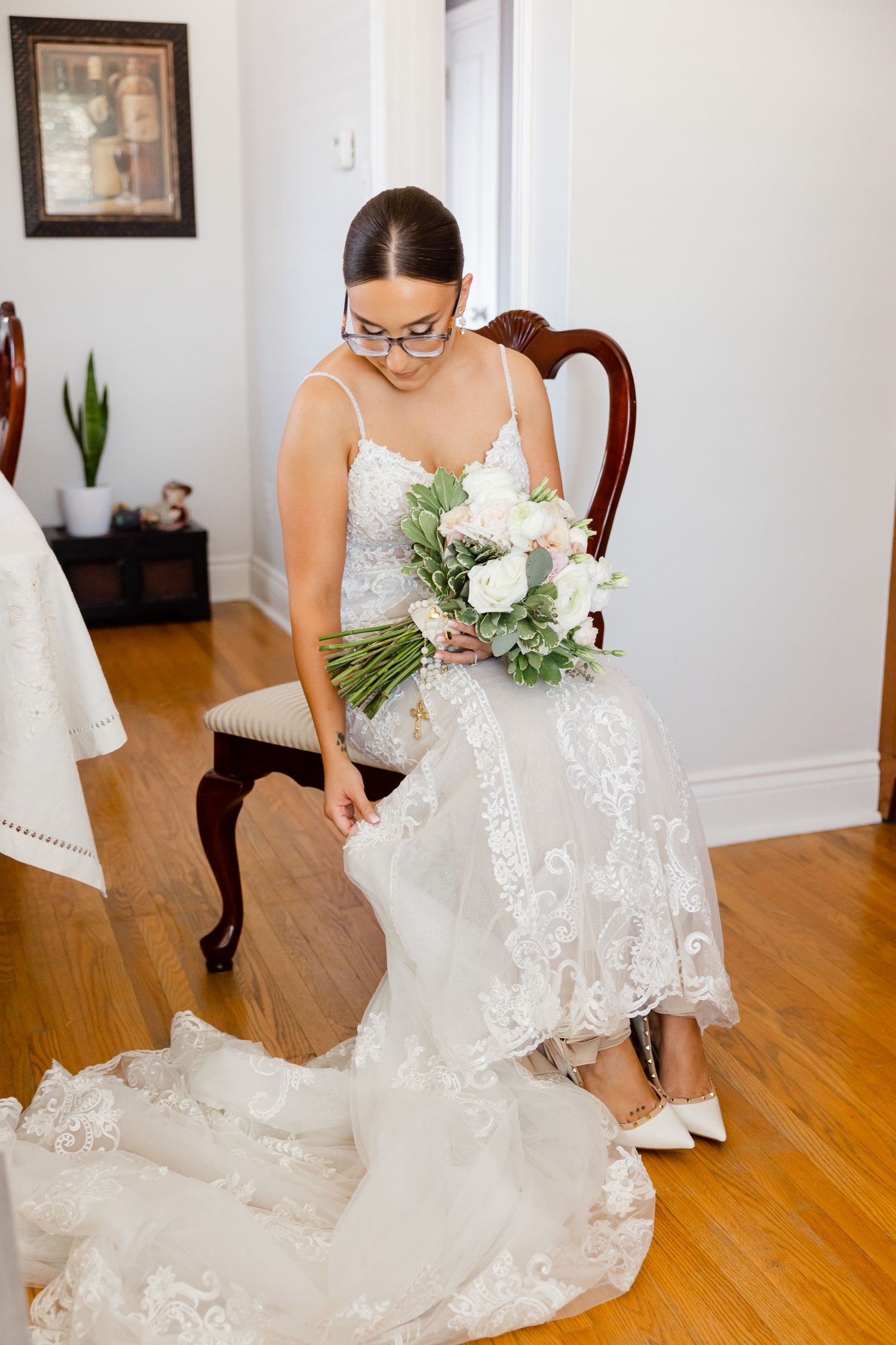 A bride in glasses adjusts her lace wedding dress while holding a white and green bouquet.