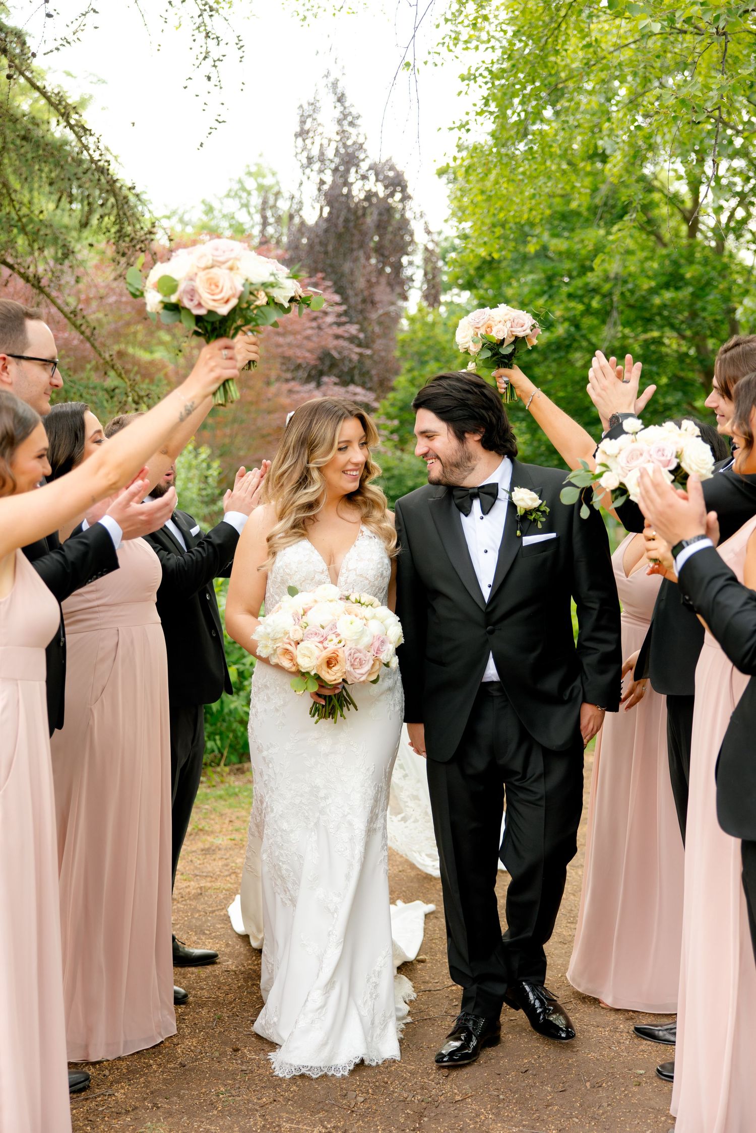 Wedding party celebrates with raised bouquets as couple walks through in garden setting.