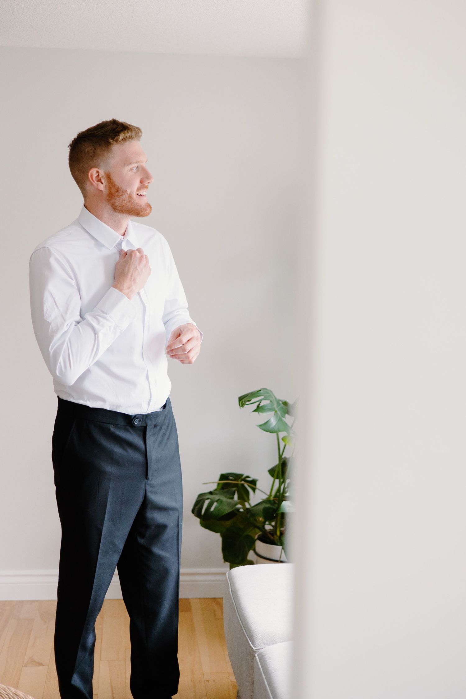 Person in white dress shirt and dark pants adjusting tie next to potted plant.