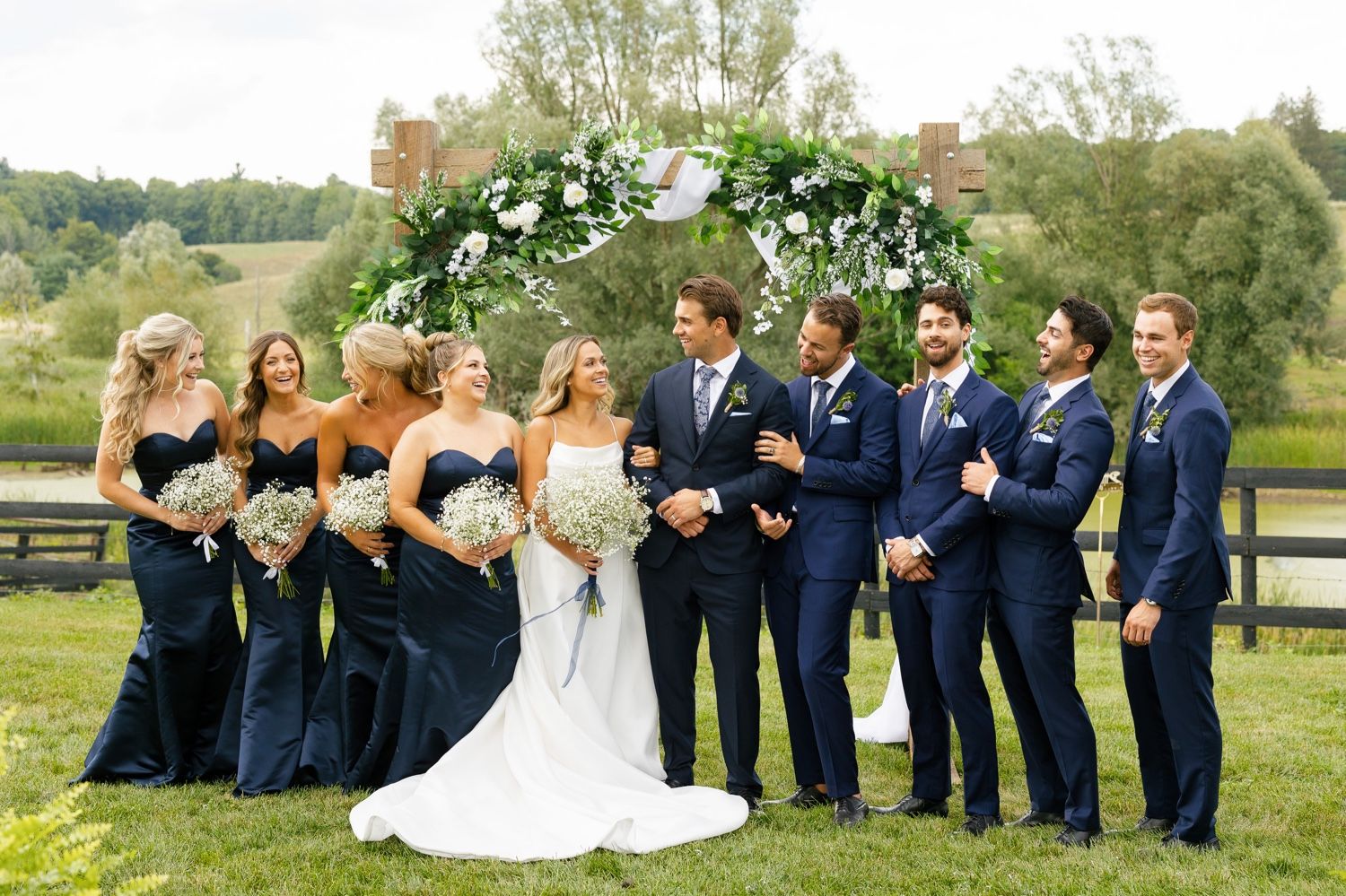 Wedding party poses under a floral arch with bridesmaids in navy dresses and groomsmen in blue suits.