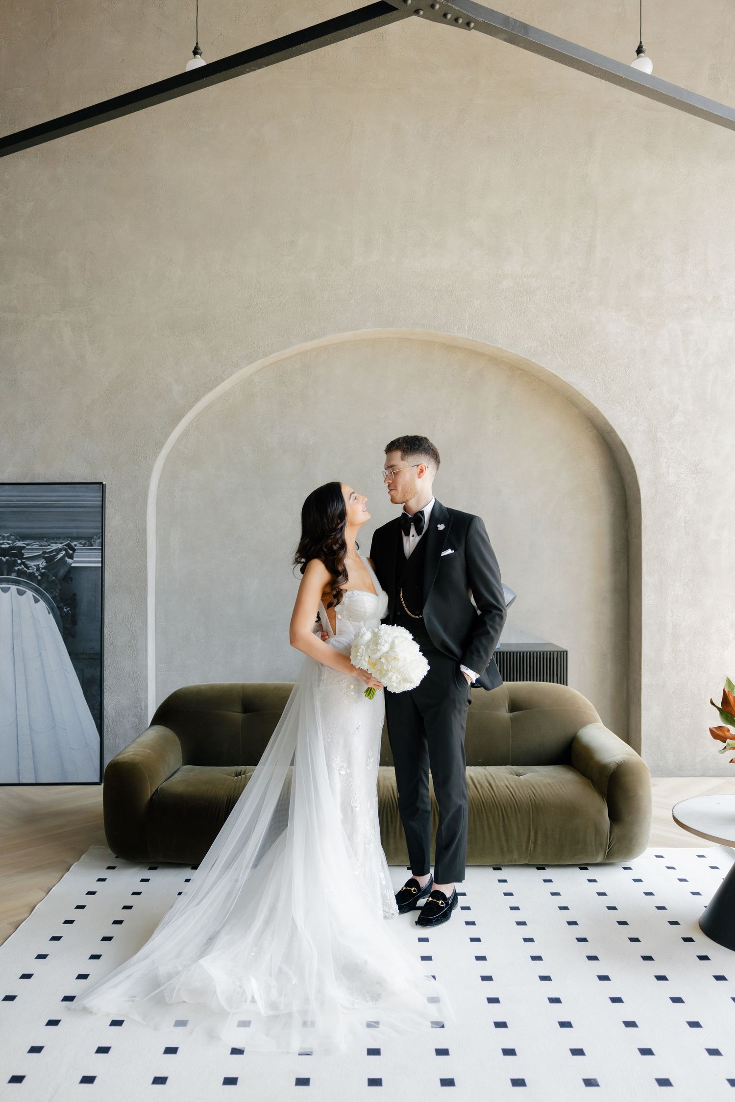 Modern wedding portrait with couple posed against minimalist arch and olive velvet couch.