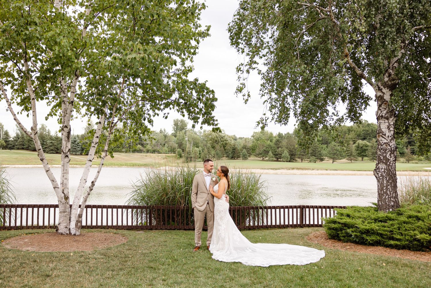 Scenic outdoor wedding photo by a lake with birch trees and wooden fence.
