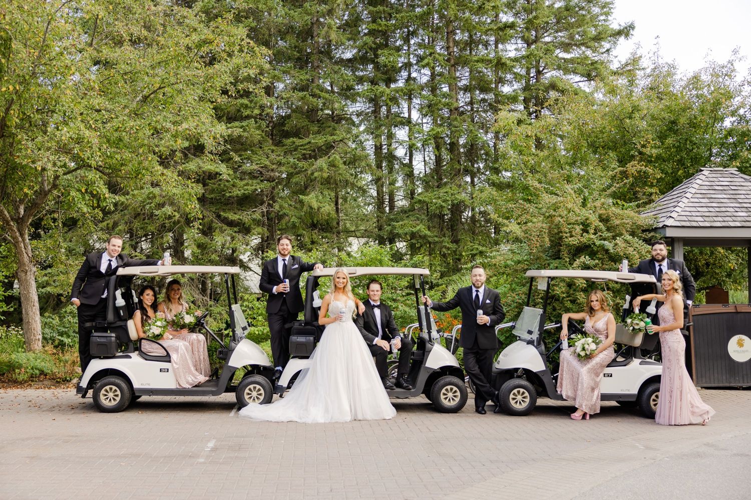 Wedding party posing with golf carts on a tree-lined path at a country club.