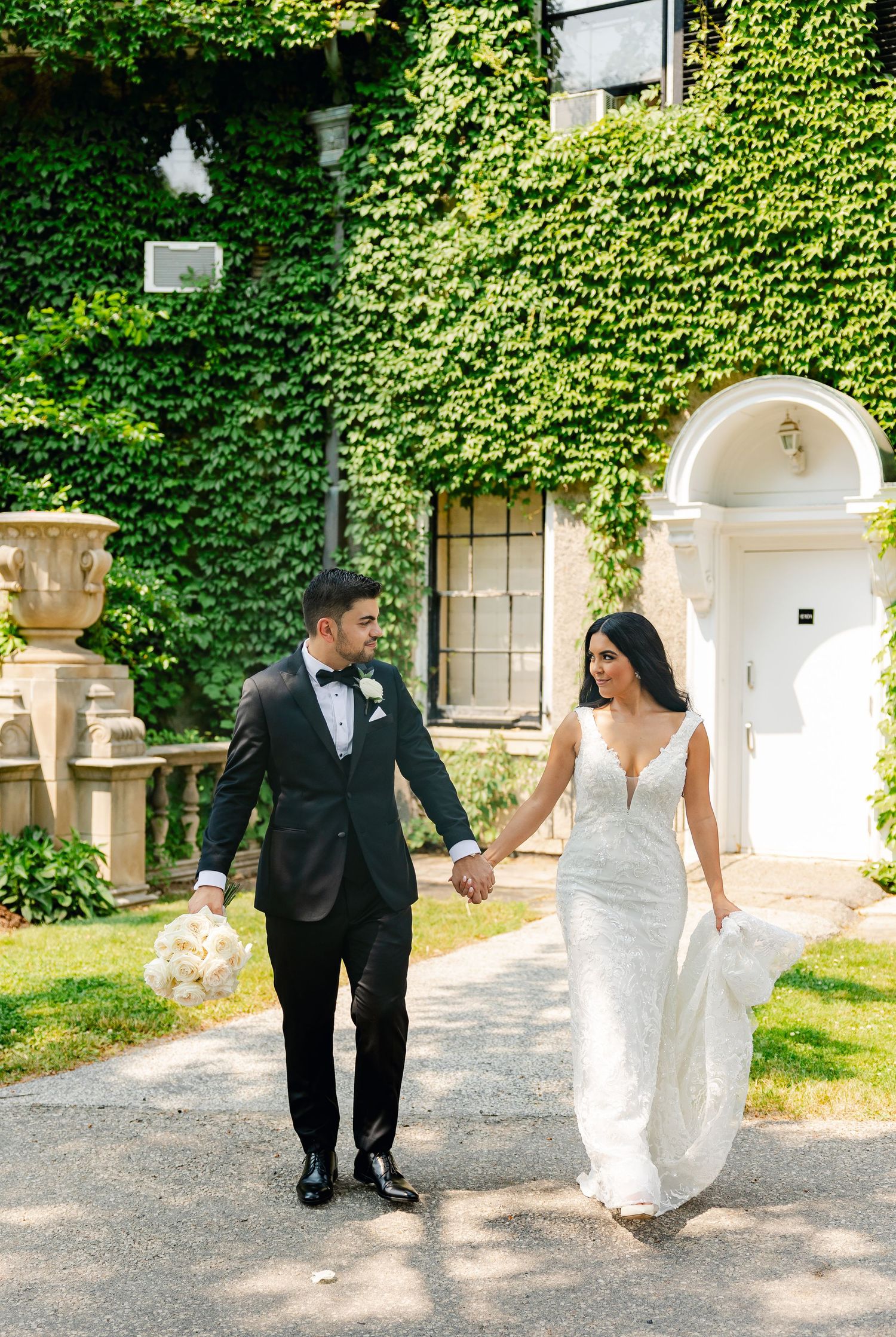 A bride in white gown and groom in tuxedo hold hands while walking along a garden path.