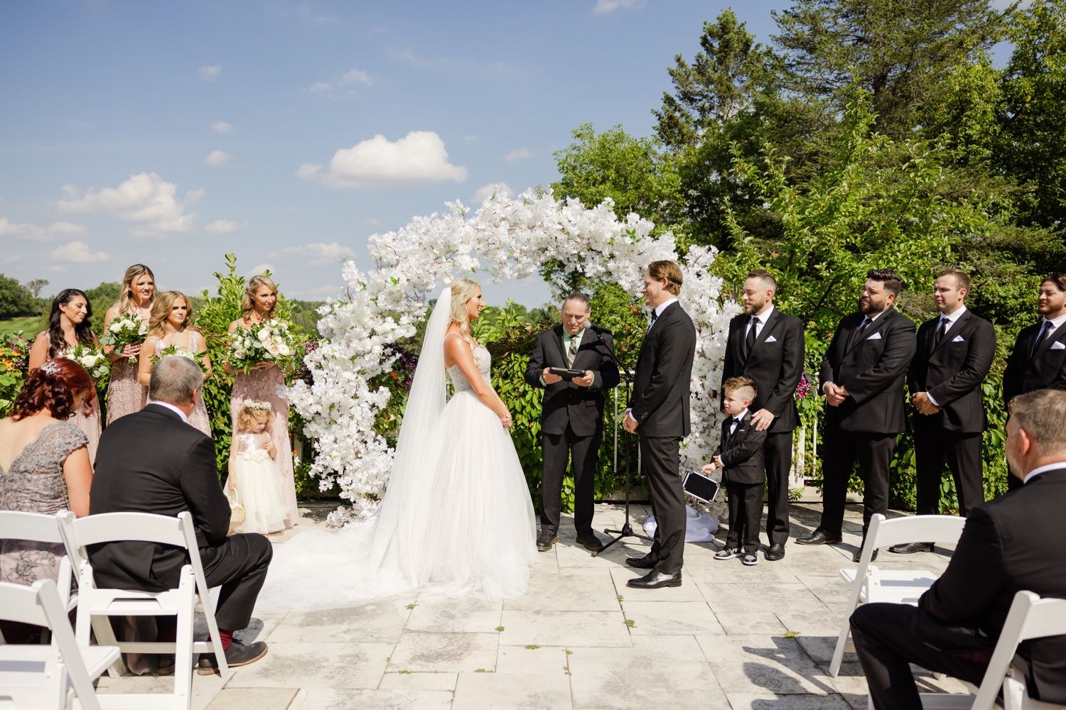 Outdoor wedding ceremony under an elegant white floral arch with guests seated on white chairs.