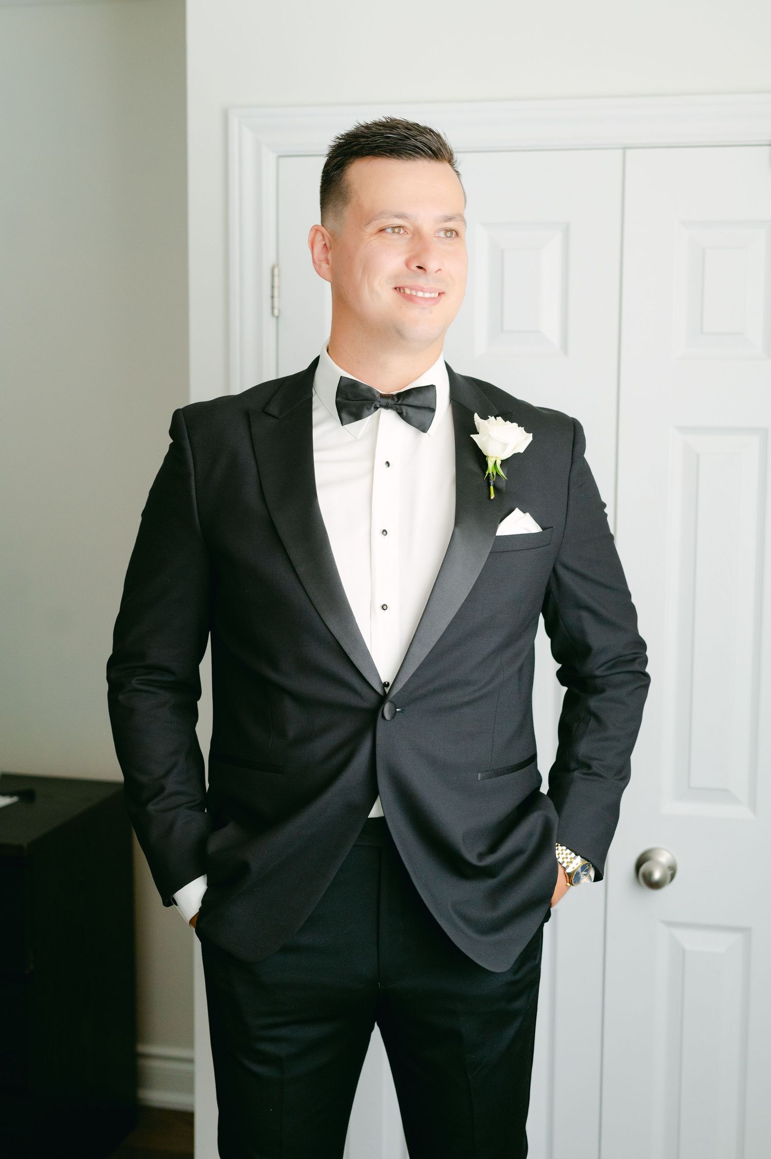 A groom poses confidently in his wedding tuxedo against a white interior backdrop.