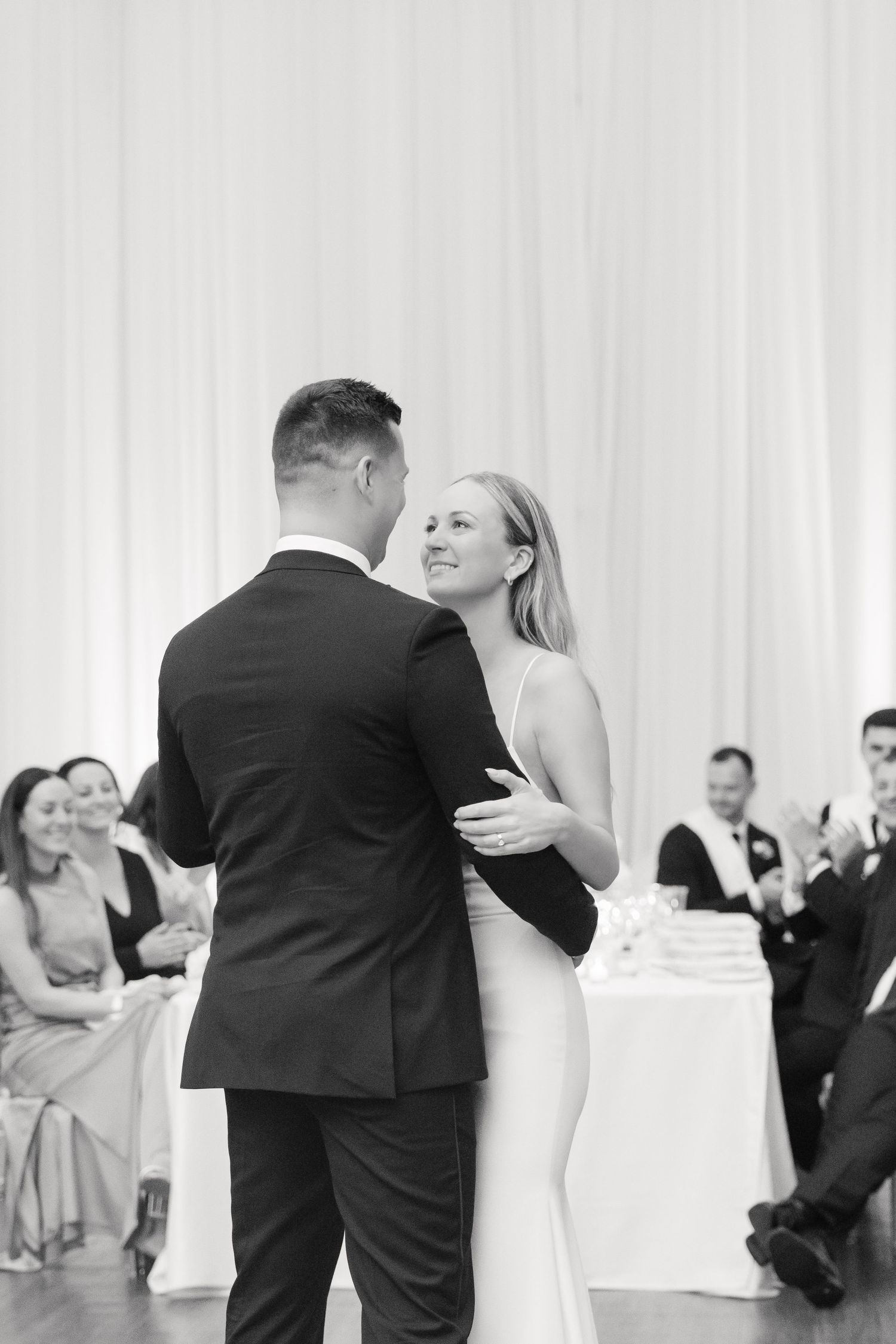 Black and white photo of a couple dancing intimately at their wedding reception with guests looking on.