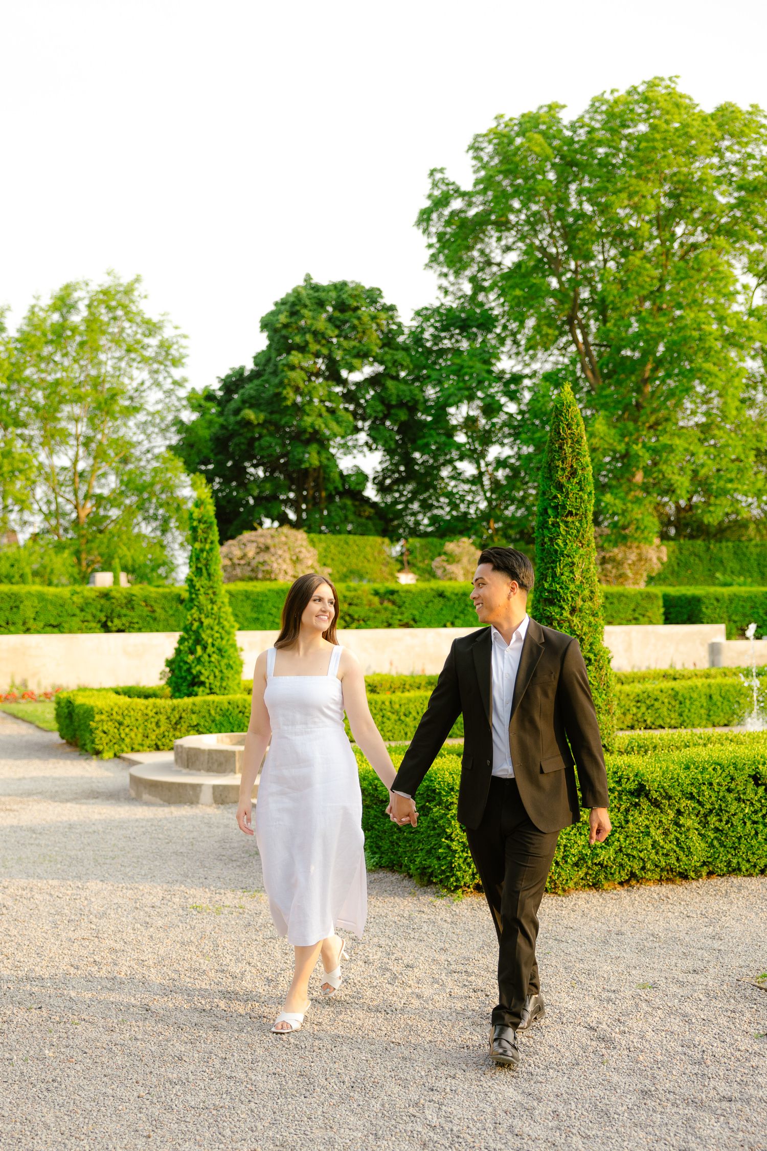 A couple takes a romantic walk on a gravel path through a beautifully landscaped garden with green topiary features.